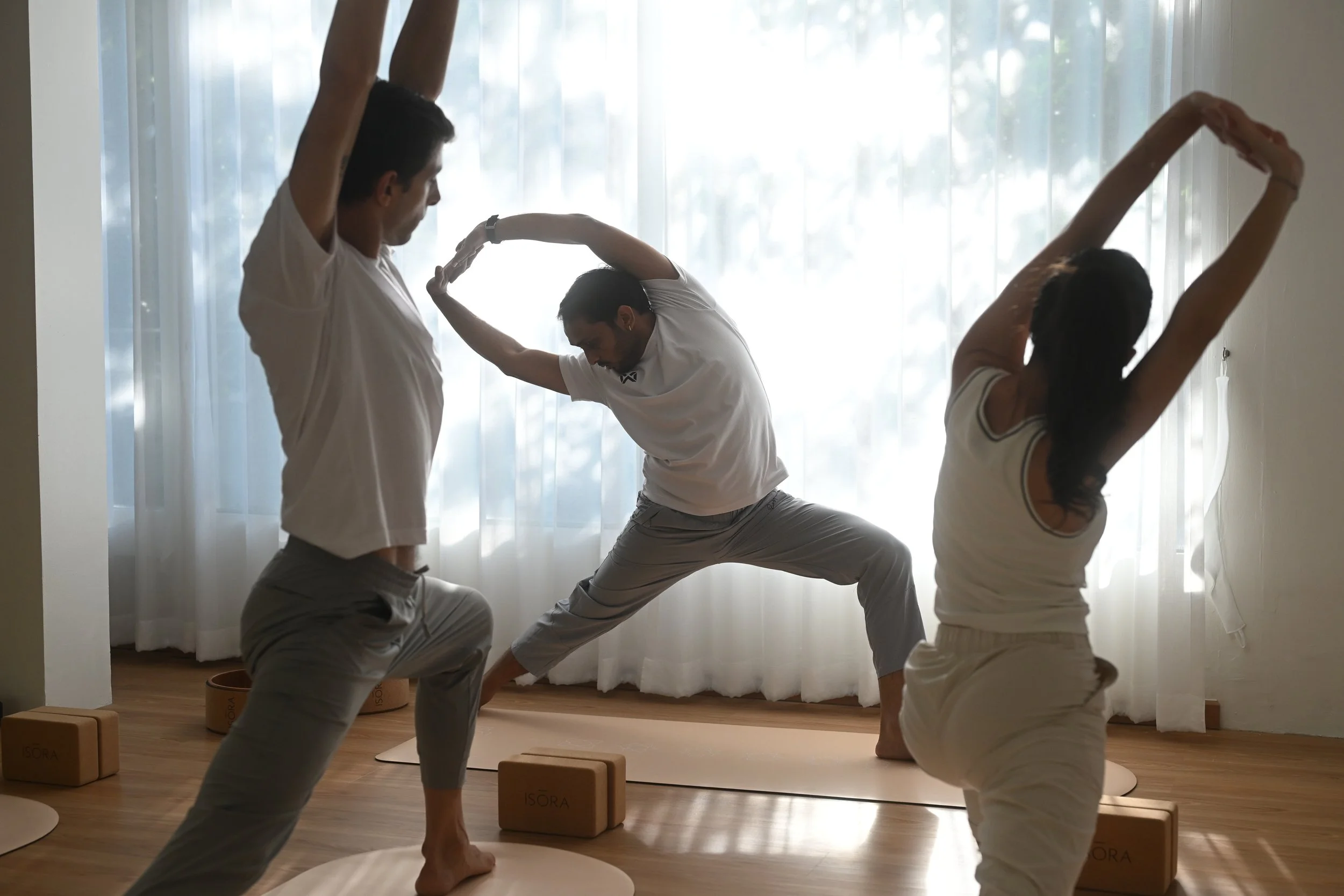 Group of people practicing yoga in a bright room with sheer curtains, performing stretching poses on yoga mats.
