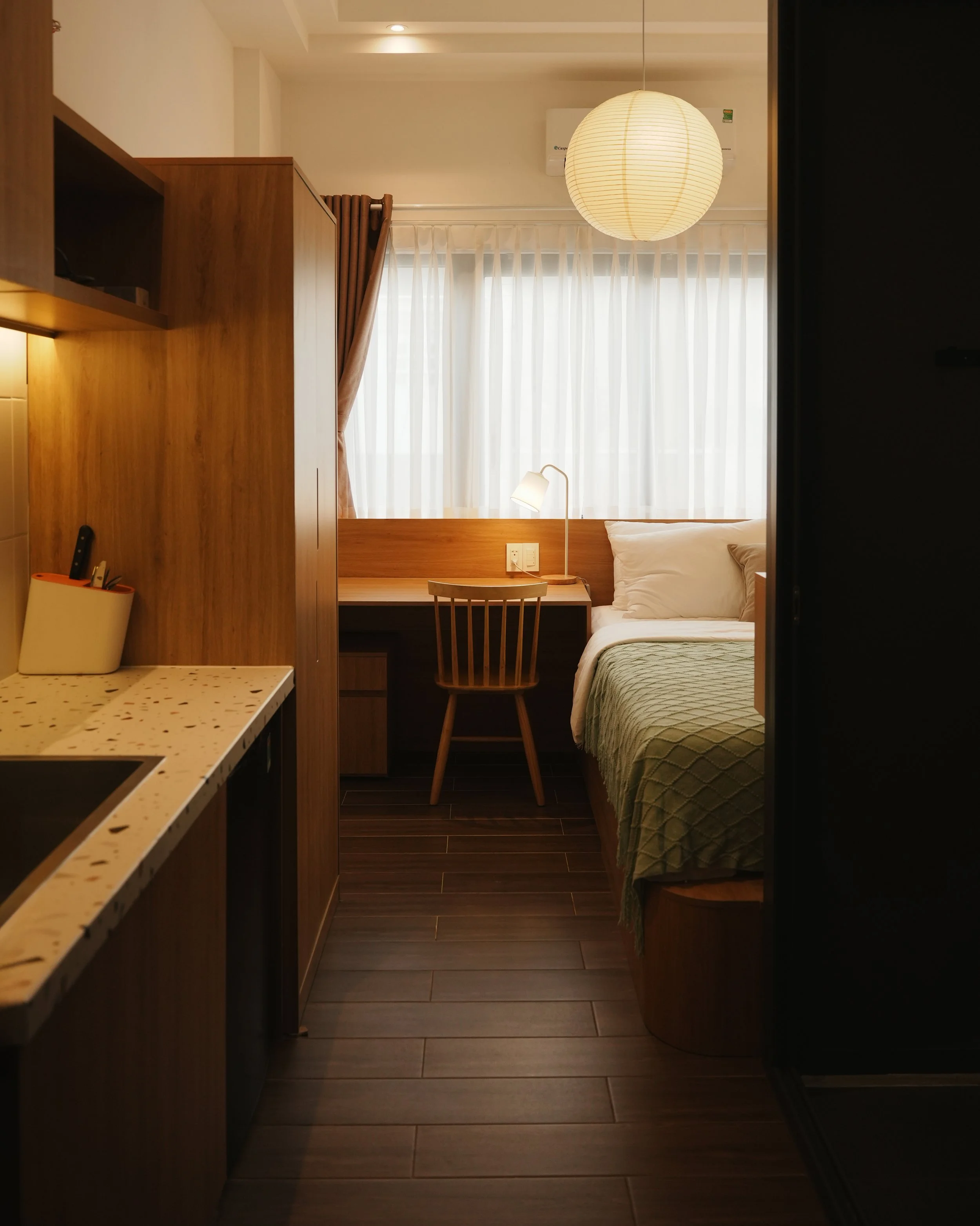 View of a small cozy hotel room with a bed, desk, chair, and window with sheer curtains, warm lighting, and wooden furniture.