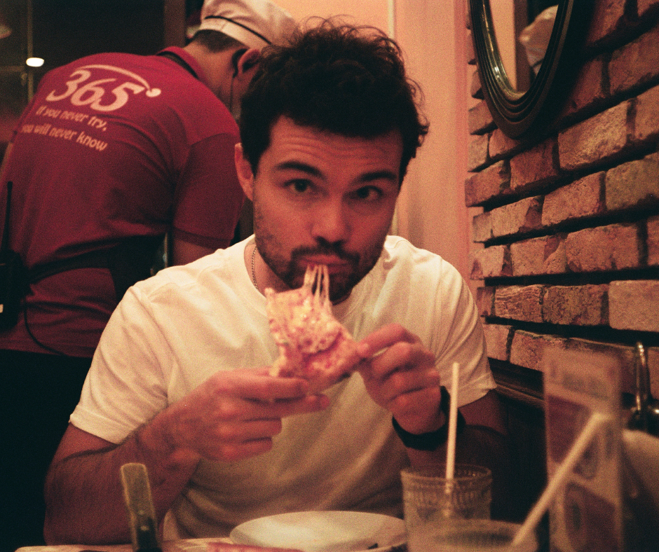 A man with dark hair and a beard eating pizza in a restaurant with brick walls, while a waiter in a red shirt is in the background.