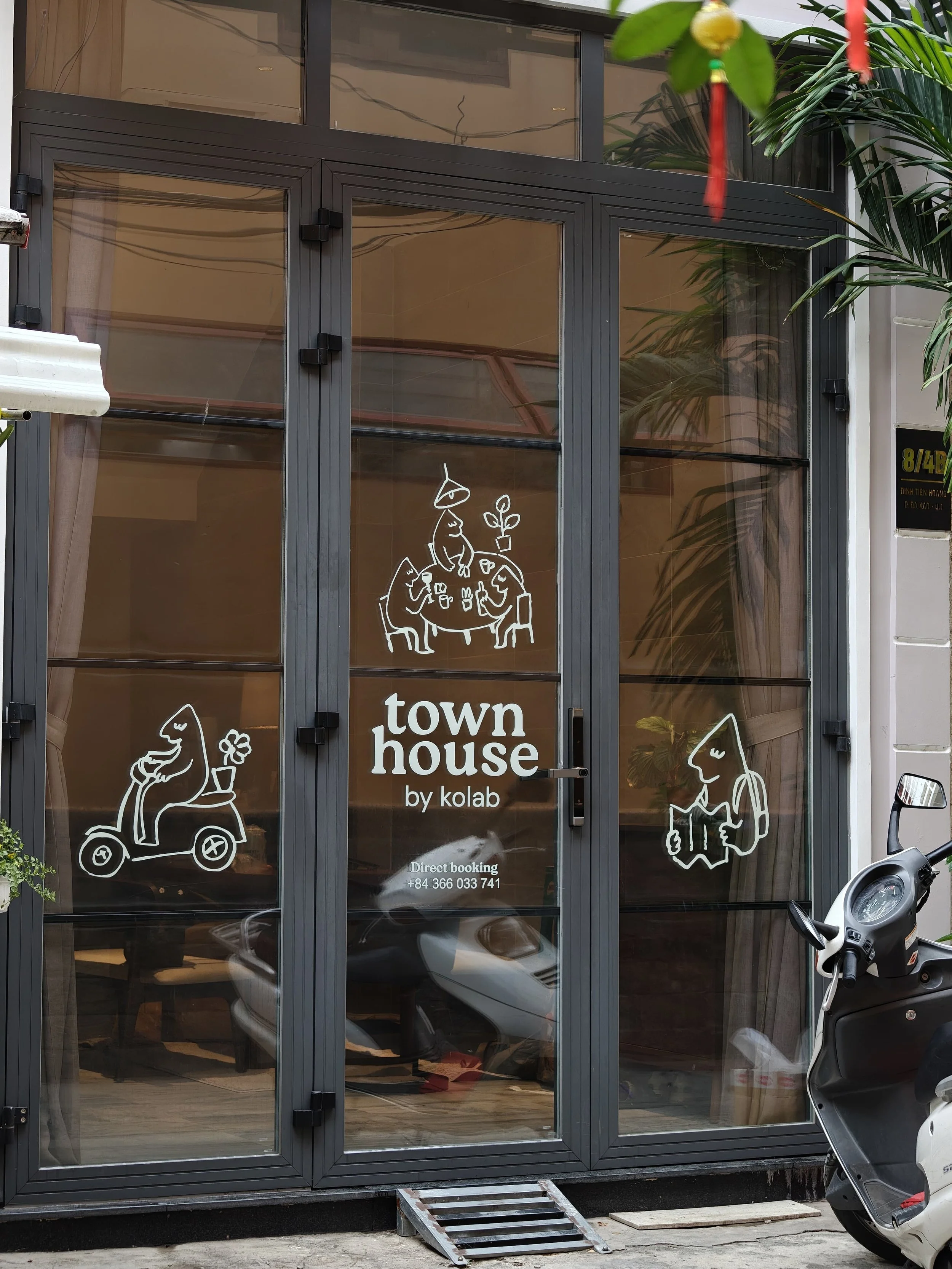 Glass storefront with illustrations of people, including a person on a scooter, two sitting at a table, and a person reading a book, along with the words 'town house by kolab' and contact information for direct booking.