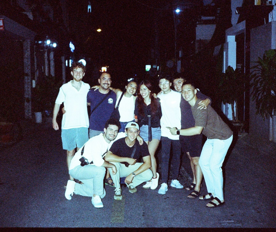 A group of nine young adults posing together on a street at night, smiling and enjoying their time.