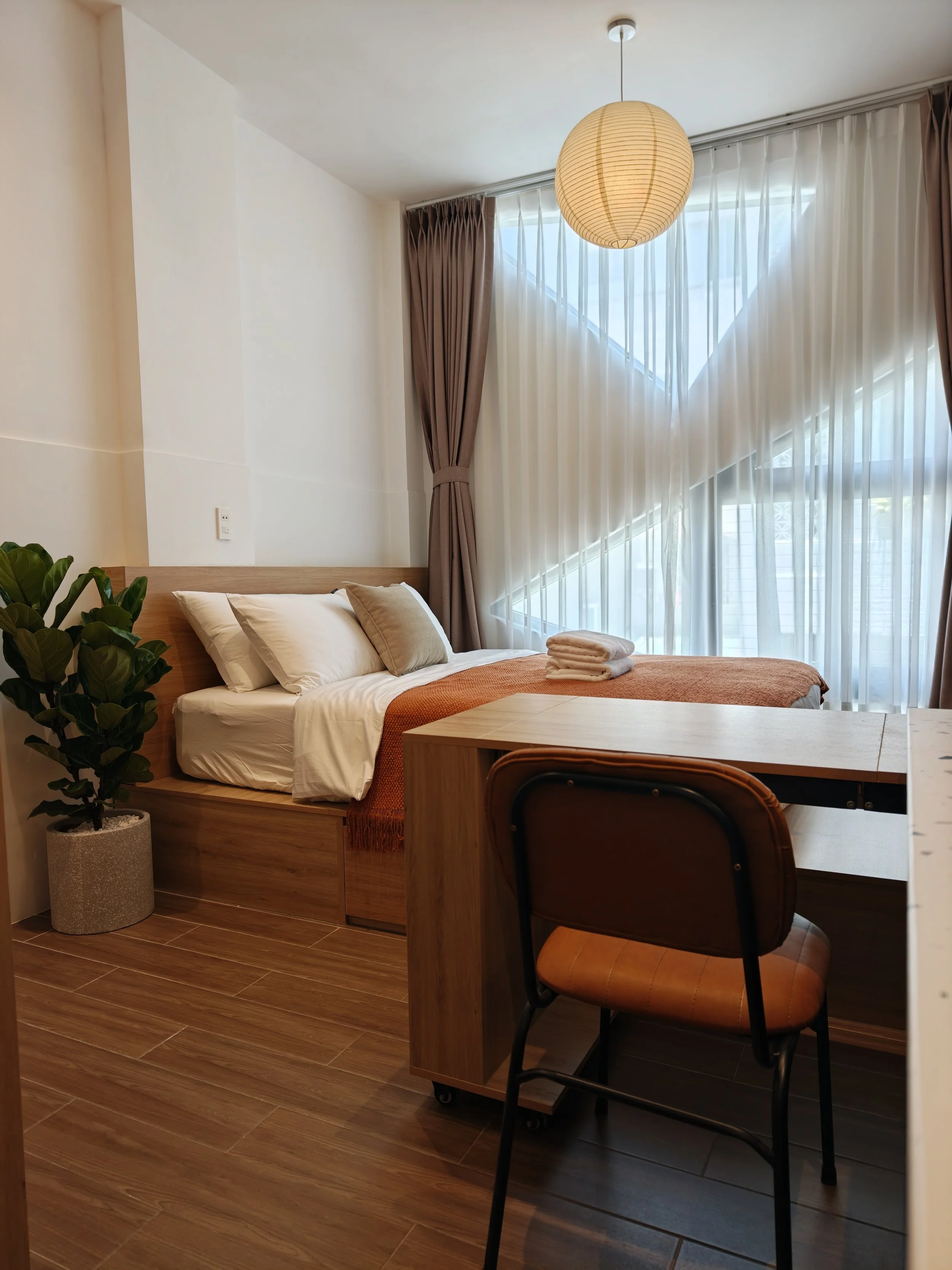 A cozy bedroom with a bed featuring white bedding, a brown blanket, and pillows, next to a potted plant. There is a wooden desk and chair, large windows with sheer white curtains and brown drapes, and a round paper lantern hanging from the ceiling.