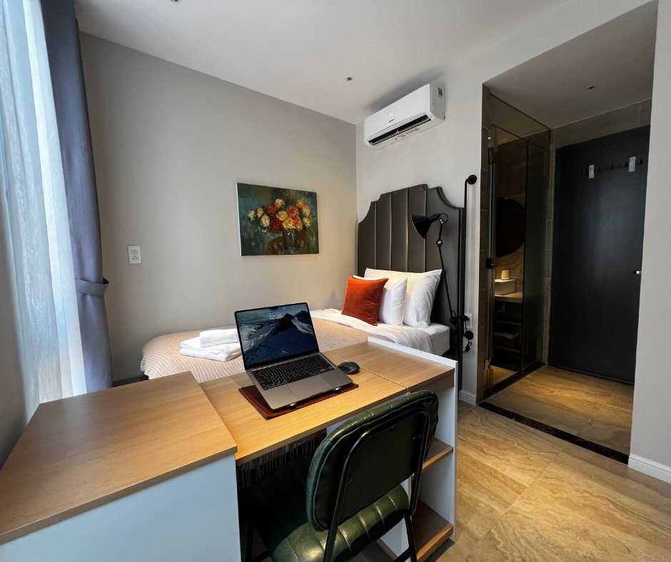 A small hotel room with a single bed, a desk with a laptop, a chair, a painting of flowers on the wall, and an air conditioning unit above the bed.