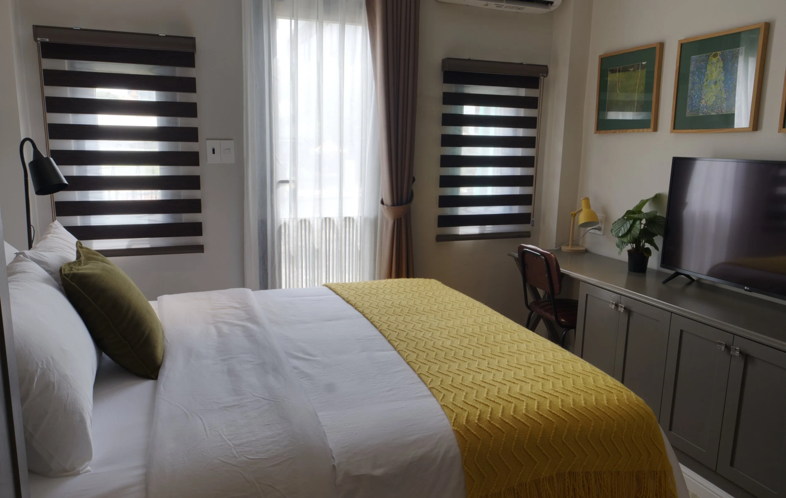 A cozy bedroom with a large bed covered with white sheets and a yellow textured blanket, accented with green pillows. There are striped blackout curtains on the windows, a gray cabinet with a flat-screen TV, a small desk with a yellow lamp and green plant, and framed artwork on the wall.