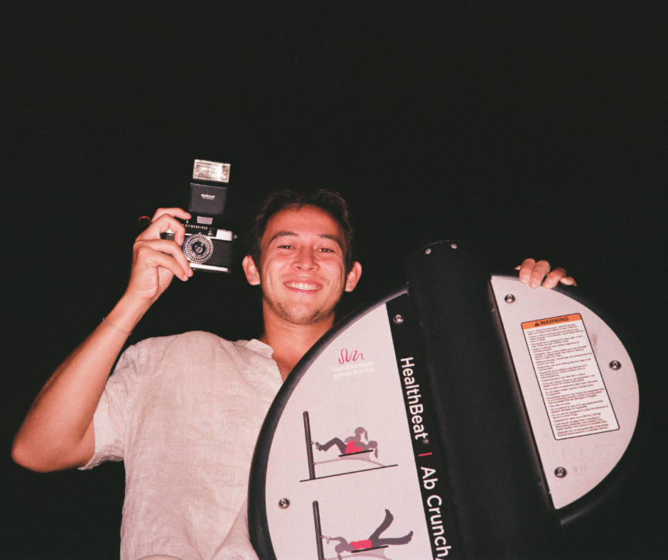 A young man smiling and holding a camera flash above his head, standing behind a fitness machine labeled 'HeathBeat | Ab Crunch,' at night.