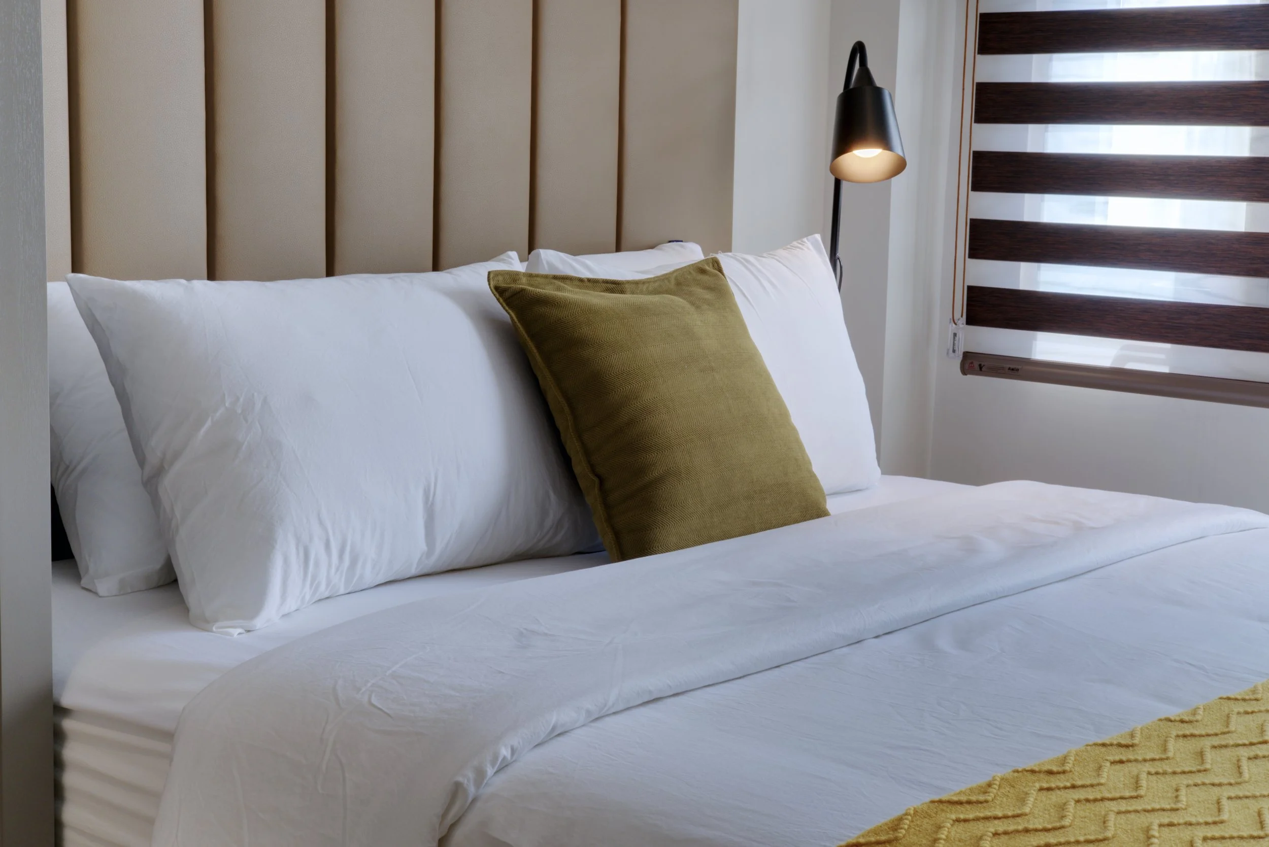 A neatly made bed with white sheets and pillows, a green accent pillow, a beige headboard, a black reading lamp, and a window with partial blinds.