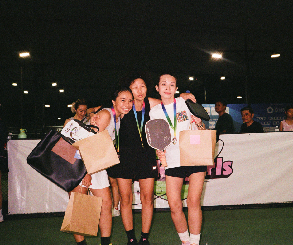 Three young women smiling and holding paddle racquets and gift bags at an indoor tennis event, with other people in the background.