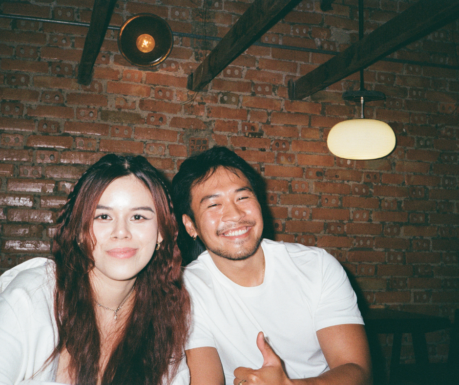 Two people, a woman and a man, sitting together smiling, in a dimly lit room with exposed brick walls and hanging pendant lights.