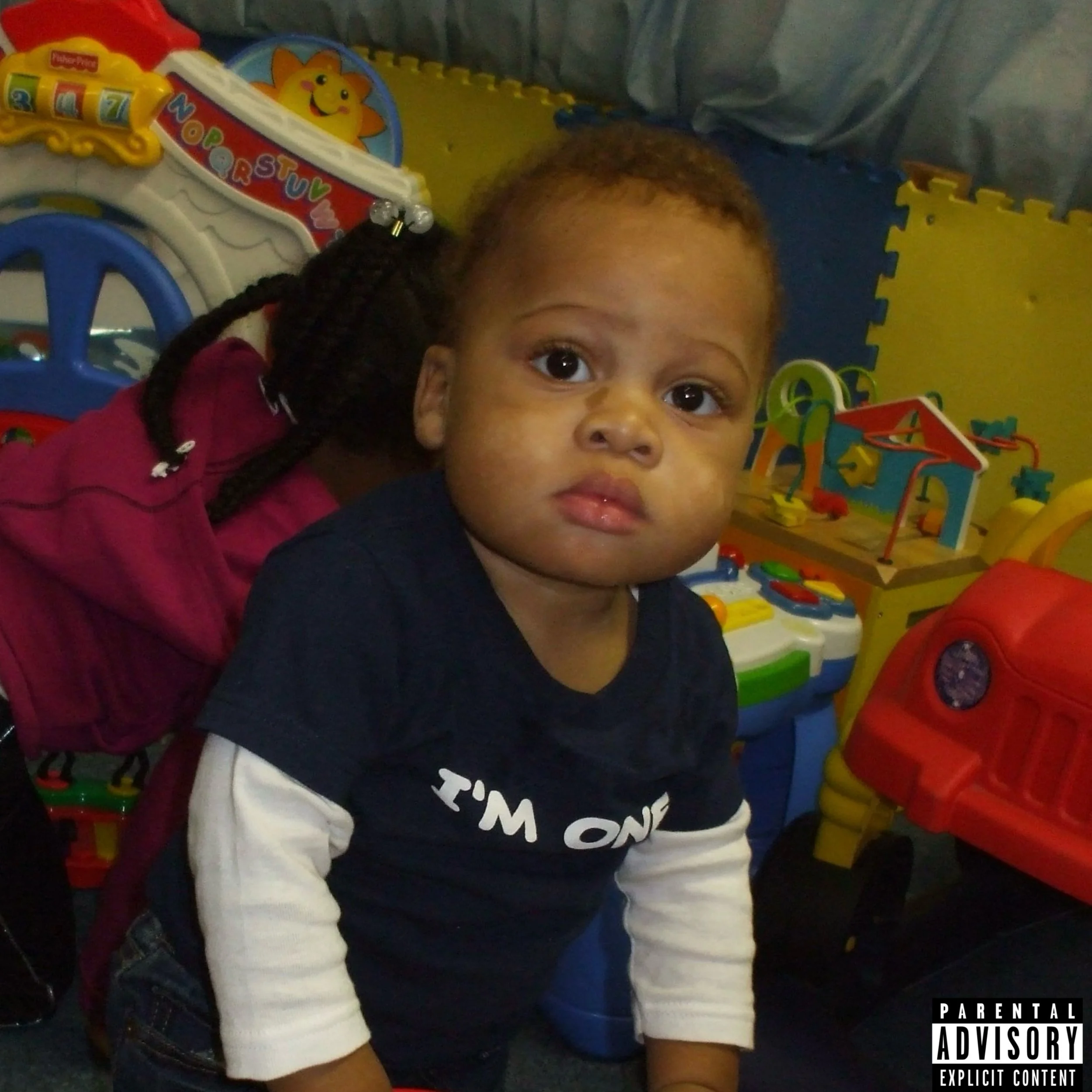 A young boy with short curly hair and brown skin looking at the camera in a playroom with toys in the background.