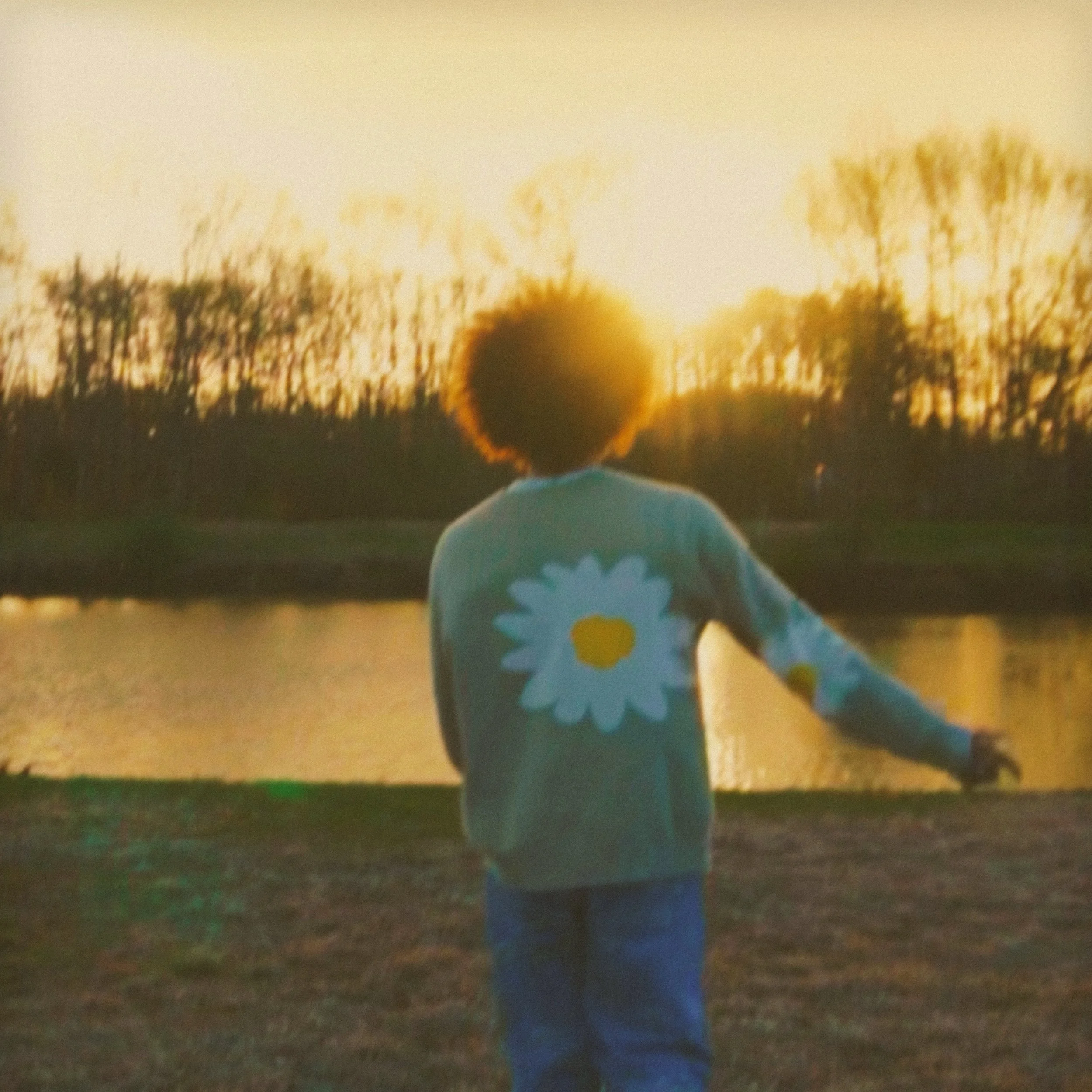 A person with curly hair wearing a gray sweater with a large white and yellow daisy on the back, standing by a body of water at sunset, with trees in the background.