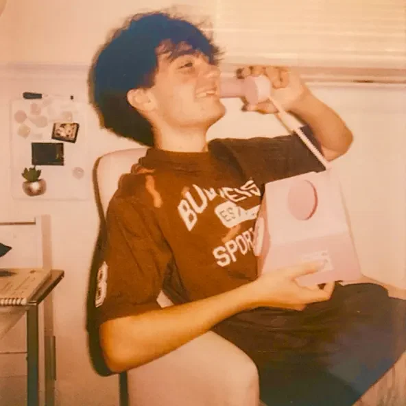 A young man with dark, curly hair smiling and talking on a pink rotary phone while sitting in a room with decorations on the wall.