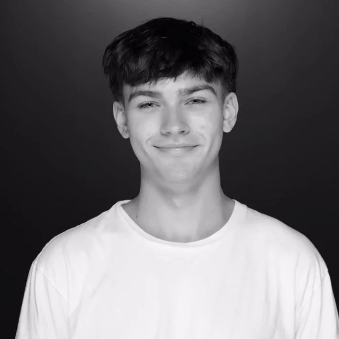 A black and white portrait photo of a young man with short dark hair, smiling, wearing a plain white T-shirt, against a dark background.