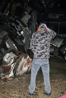 Person wearing a camo jacket and jeans, standing outdoors at night, facing broken and wrecked cars.