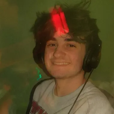 A young man with headphones smiling in a dimly lit room with green and red lighting.