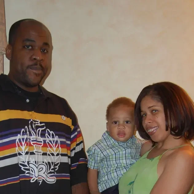 Three people, a man, a woman, and a young child, are posing indoors against a plain wall background.