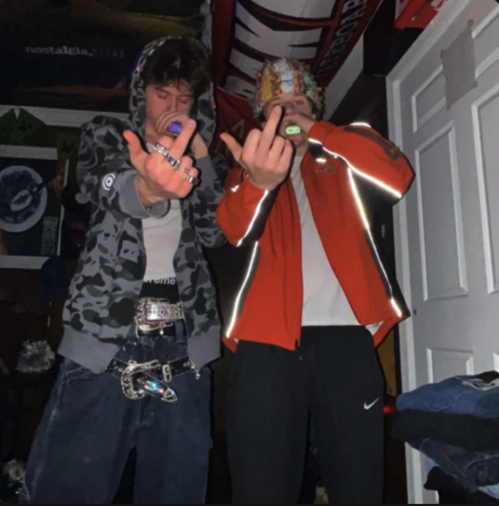 Two young men standing inside a room, both making a rude gesture with middle fingers. The man on the left has dark hair, wearing a hooded camouflage jacket, a white shirt, rings, and belts with shiny accessories. The man on the right is wearing a colorful knit hat, an orange jacket with reflective details, and black Nike sweatpants. Both are holding electronic devices near their mouths.