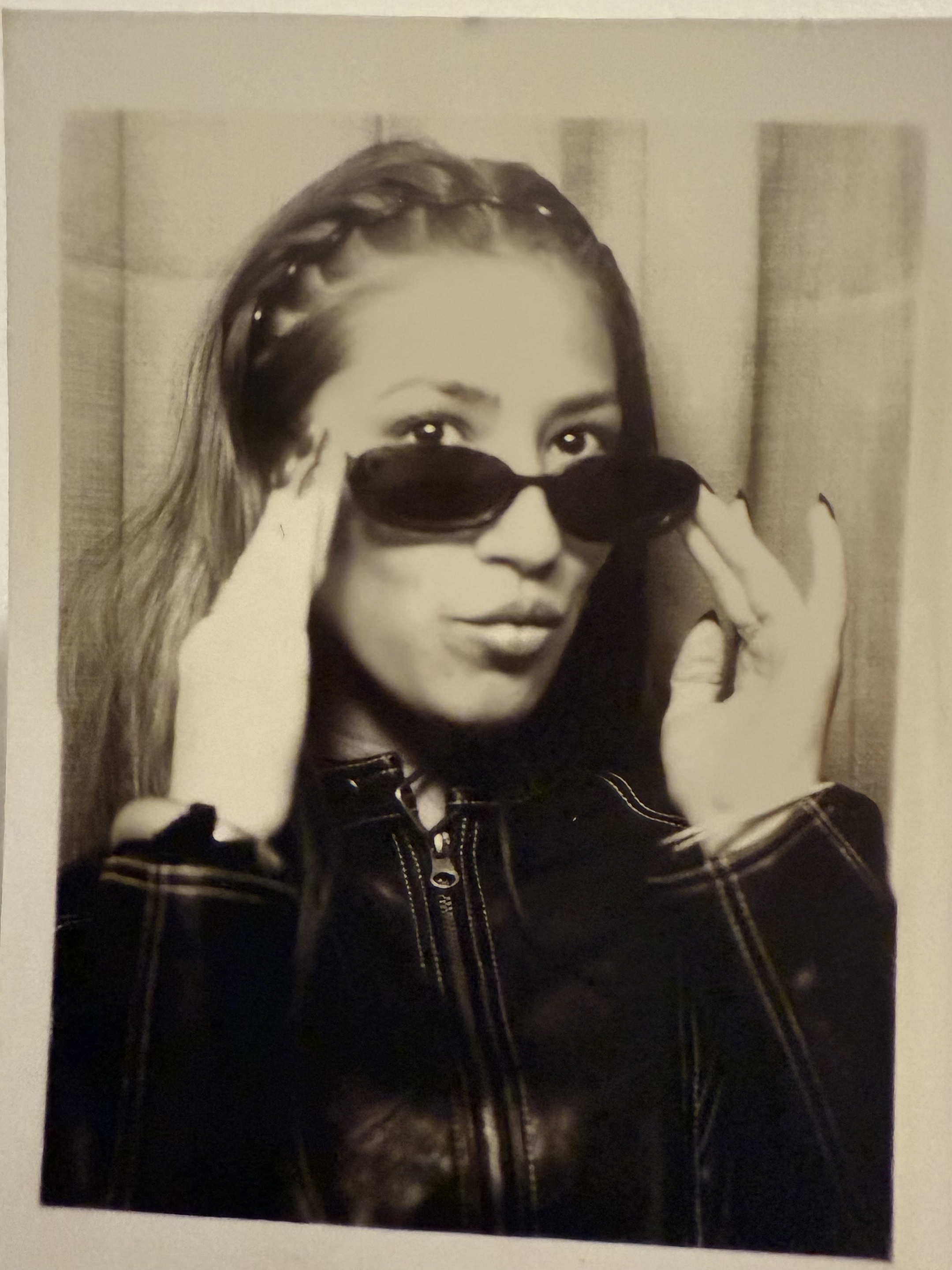Black and white photo of a young woman with long hair, wearing dark sunglasses, a leather jacket, and holding the sunglasses with both hands, looking at the camera.