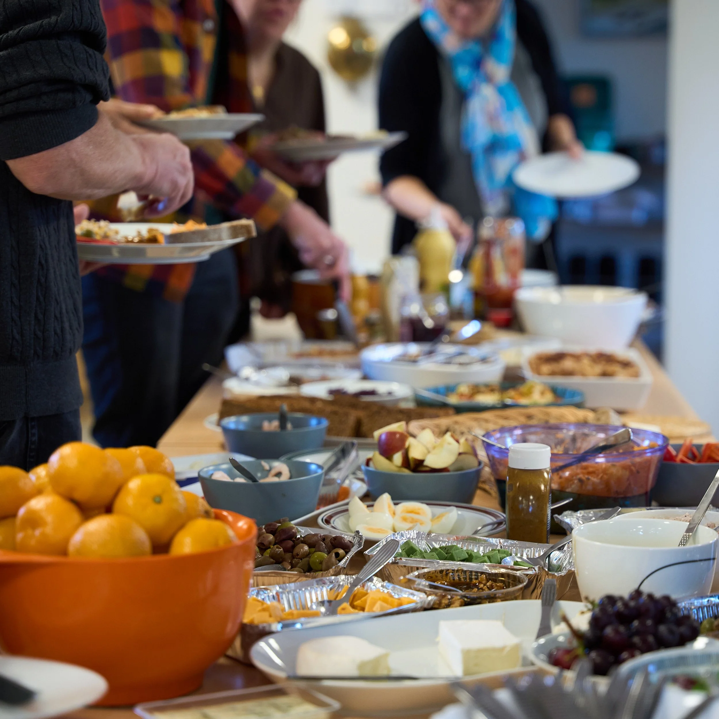 Fra vores kirkefrokost ti len søndagsgudstjeneste. Buffeetbord med forskellige retter og drikkevarer, folk står i baggrunden ved et morgenmadsbord, der er frugt, ost, pølse, kage og andre spiser.
