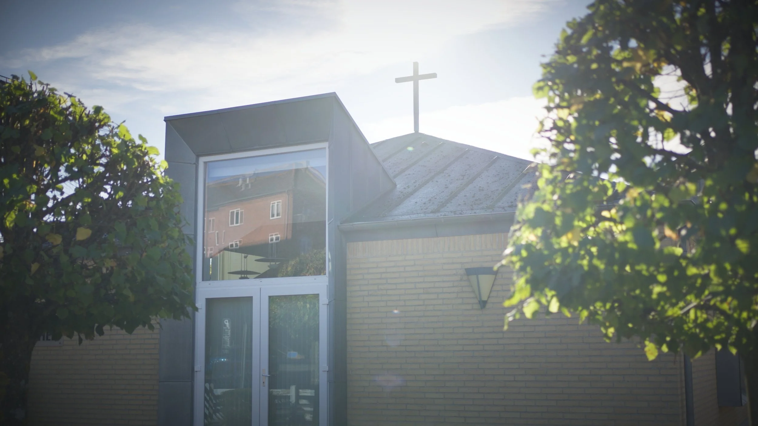 Fjordlandskirken set udefra. Der er kors på taget, omgivet af grønne træer, under en klar himmel med solskin.