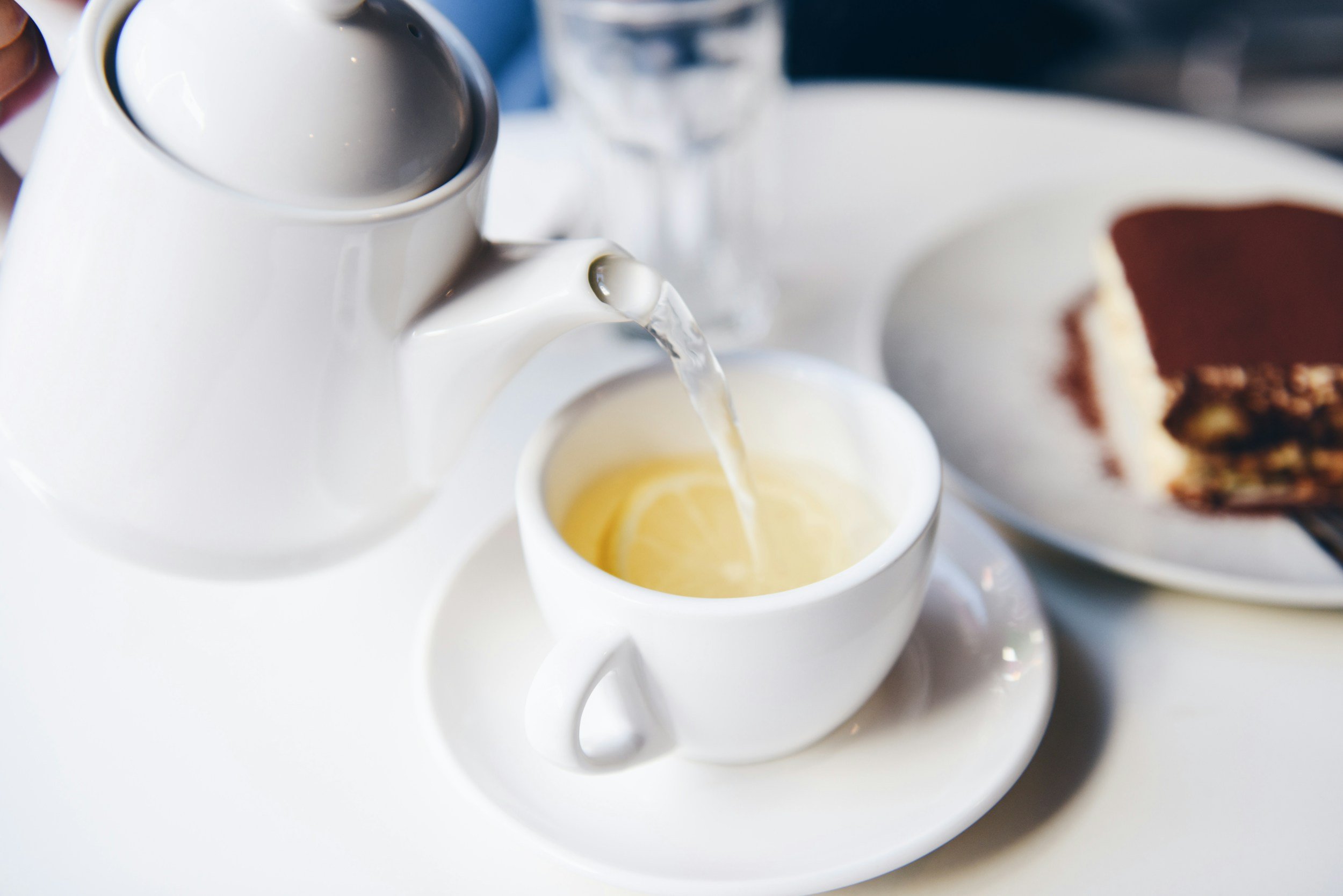 Une tasse de thé avec un citron, une théière blanche versant de l'eau chaude et un gâteau au chocolat et à la vanille sur une assiette.
