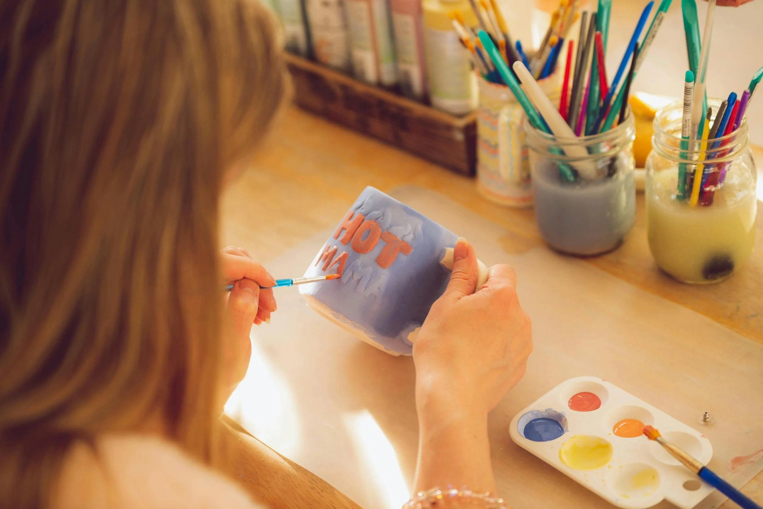Une personne peint un mug en céramique avec des lettres en couleur orange. Sur la table, il y a une palette de peinture avec des couleurs vives et plusieurs pinceaux dans des bocaux.