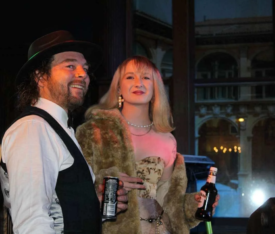 A man with a beard wearing a black hat, white shirt, and black vest smiling, and a woman with blonde hair wearing a fur coat and jewelry holding a beer bottle, at a party or social gathering indoors with a window in the background.