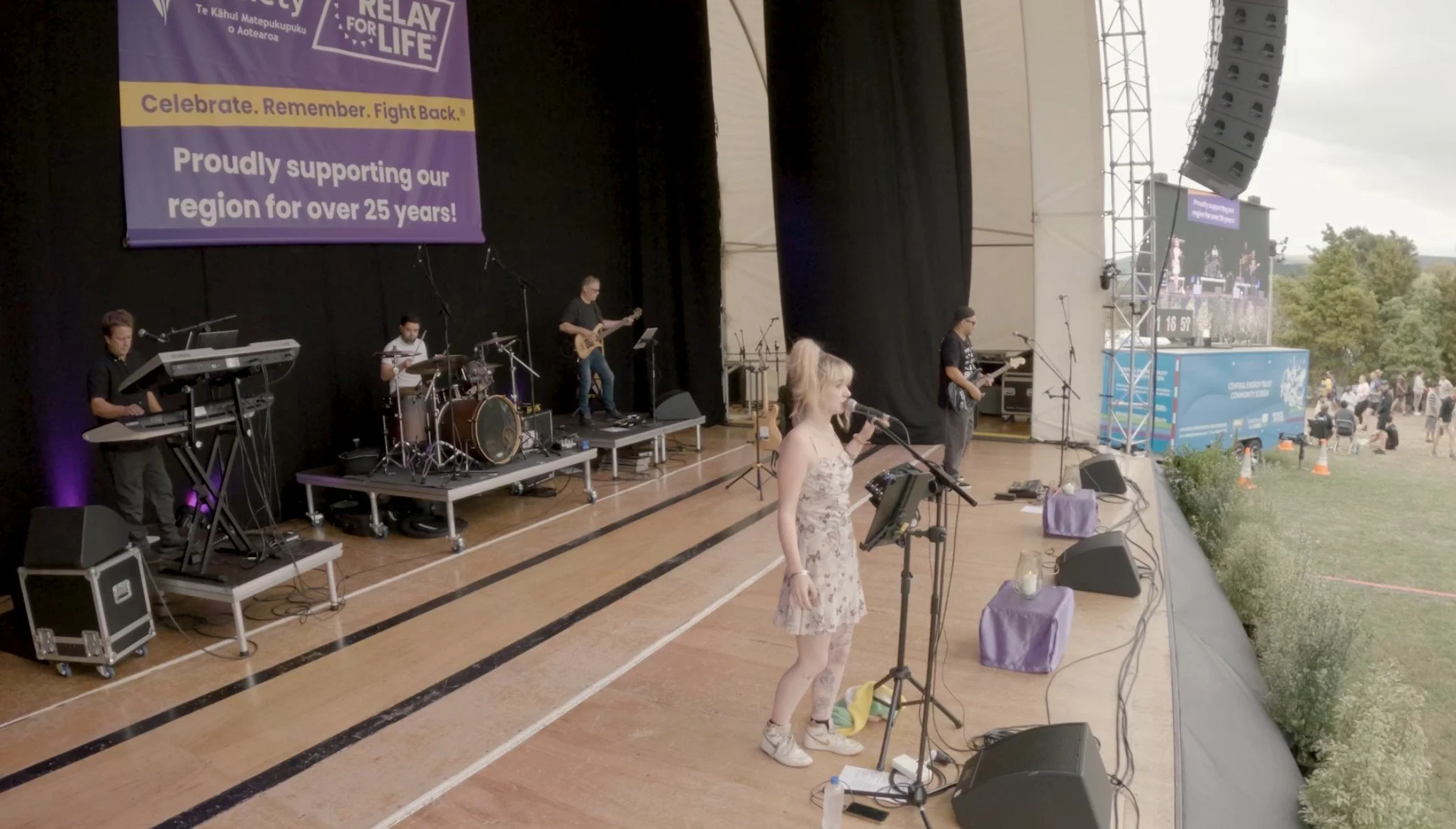 A band performs on a stage at an outdoor event with a banner supporting the Relay for Life cancer awareness fundraiser, featuring people in the background on the field.