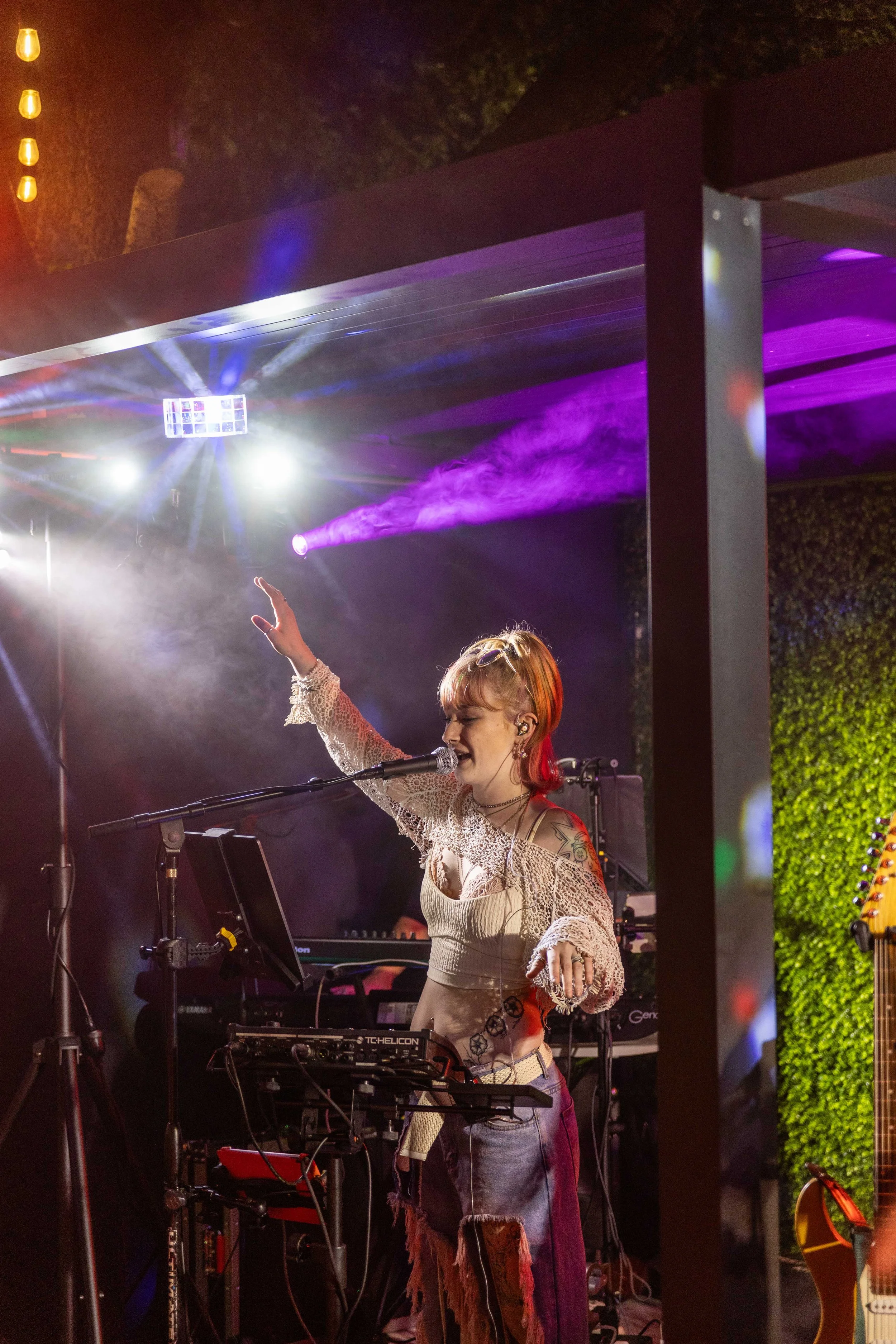 A woman singer performing on stage with colorful lights and fog effects, wearing a lace top and distressed jeans, with musical equipment around her.