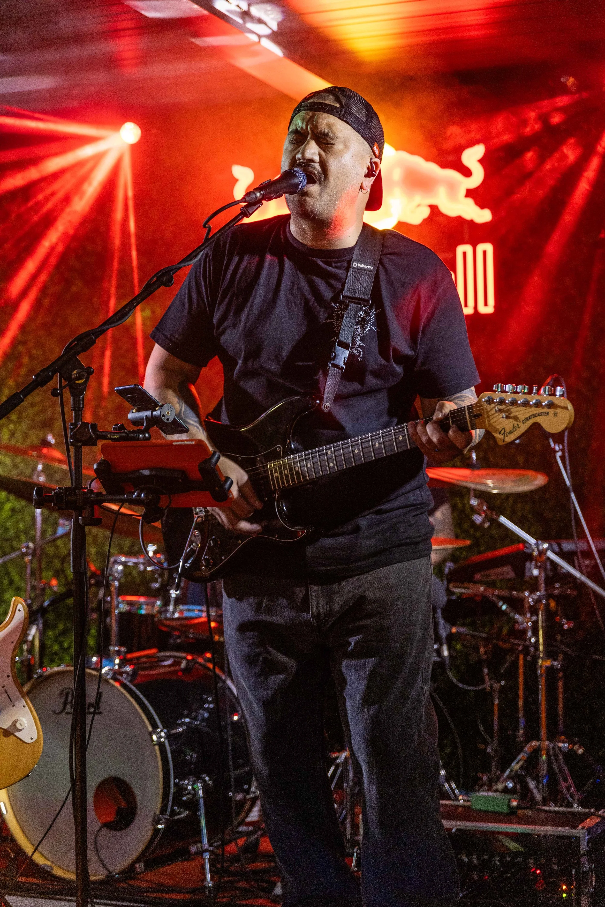 A male musician performing on stage with a guitar, singing into a microphone, with red lighting and a Red Bull logo in the background.