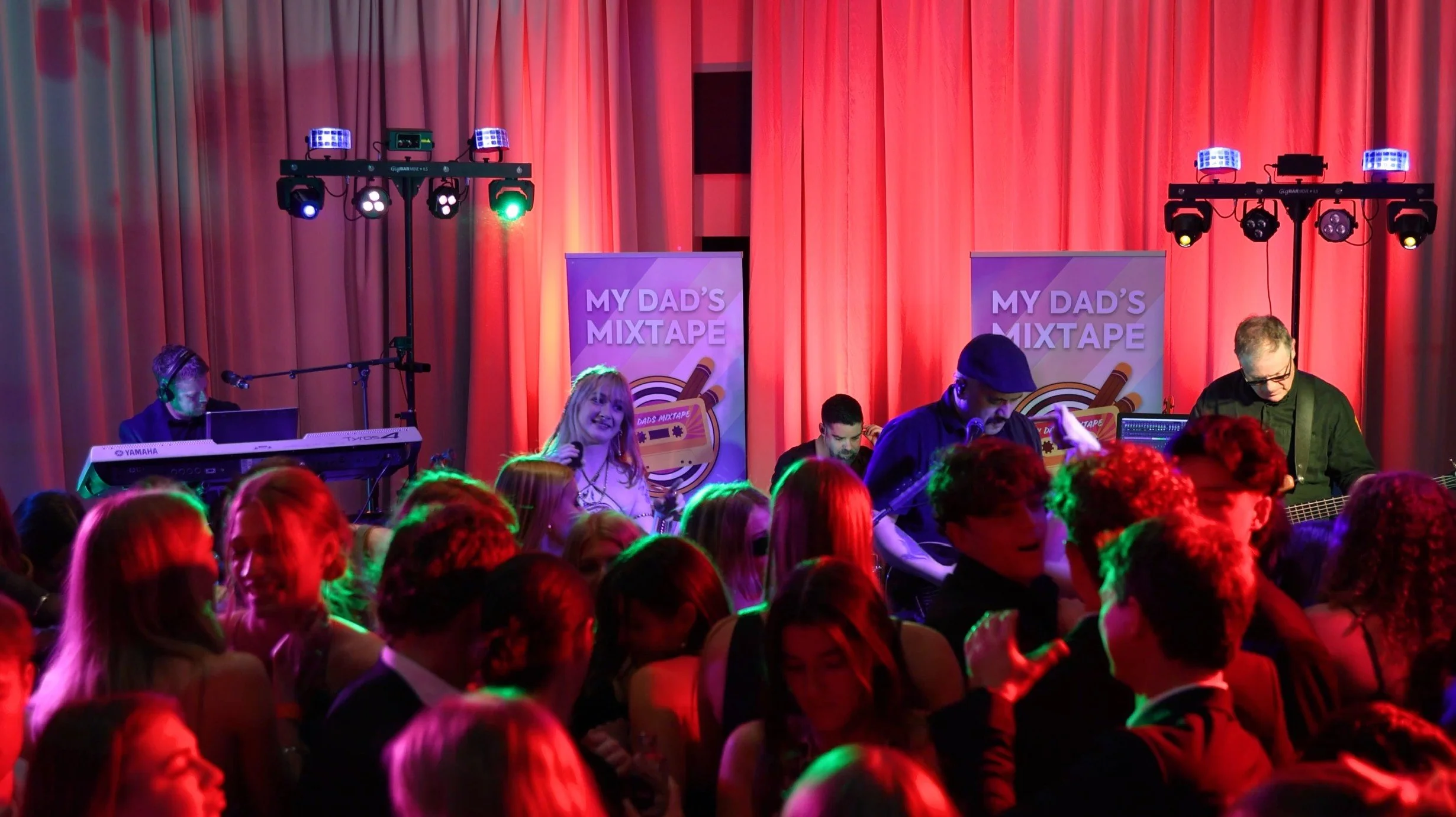 A lively crowd watching a live music performance on stage with band members and colorful stage lights, under a red curtain backdrop, with two banners reading 'My Dad's Mixtape'.