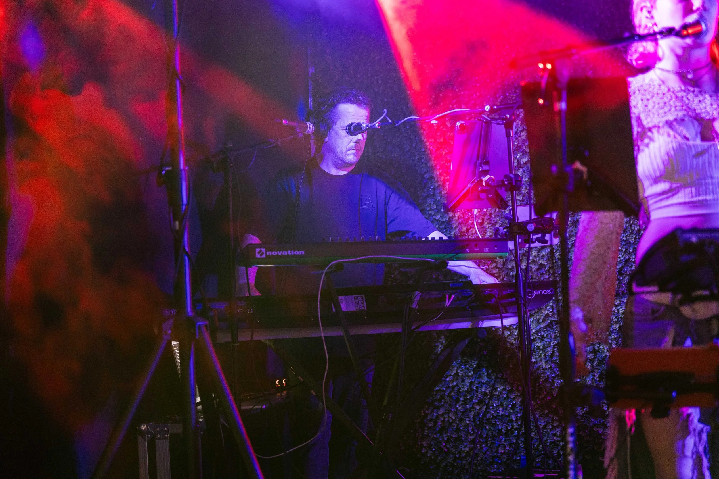 A musician playing a keyboard on stage with colorful purple and red lighting, wearing a black shirt and headphones.
