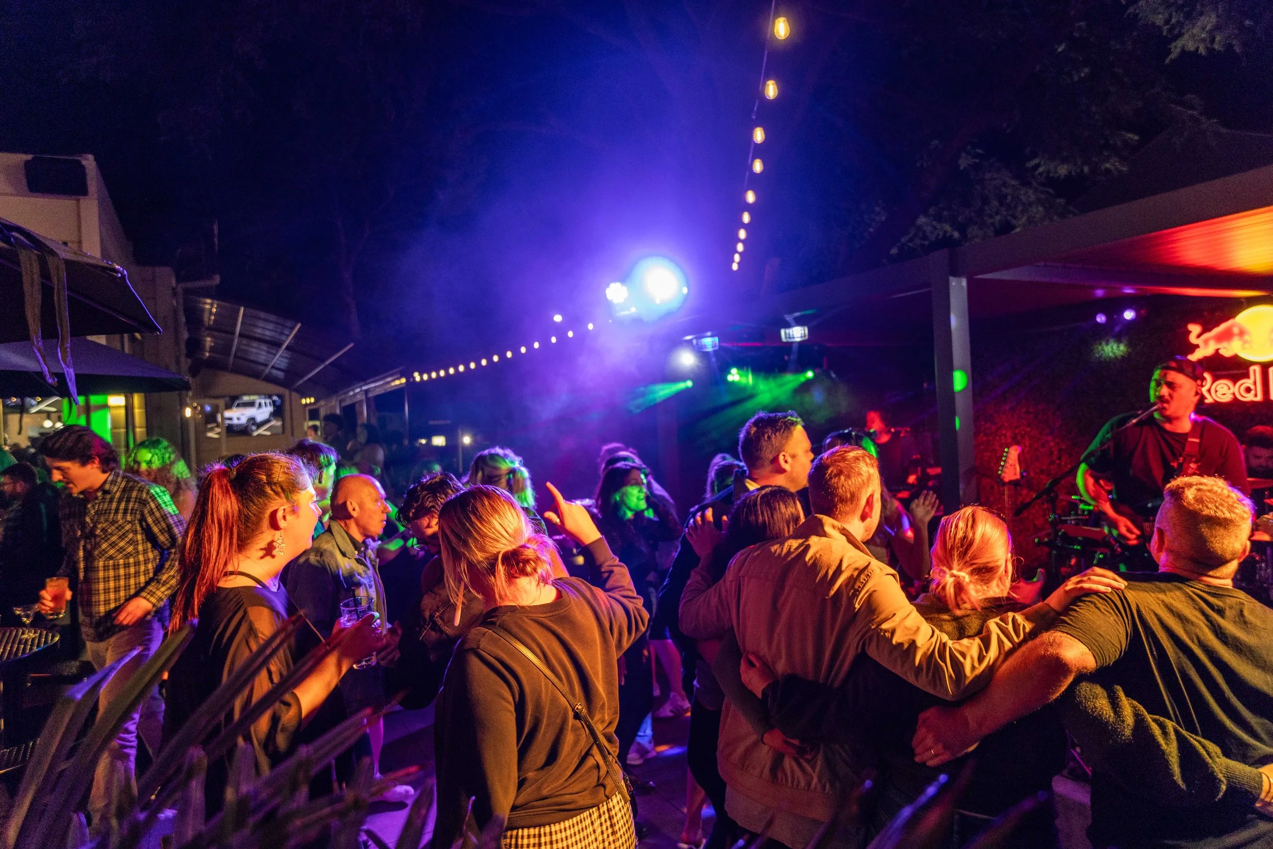 People dancing and enjoying live music at an outdoor night concert with colorful stage lights and a Red Bull sign.