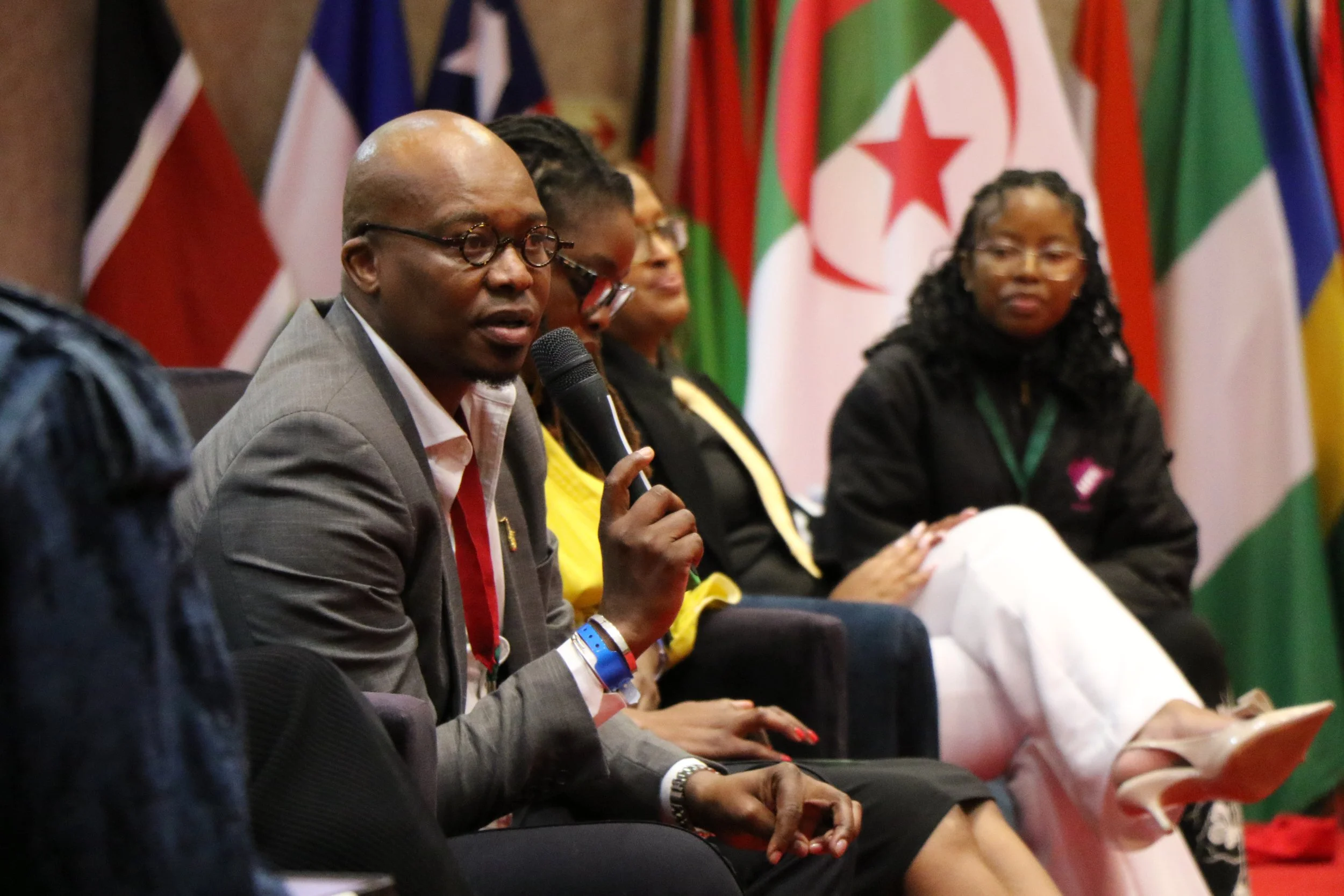 Monde Twala speaking into a microphone at a panel discussion at the AU's APRM Youth Symposium, with women seated beside him, and various African flags in the background.
