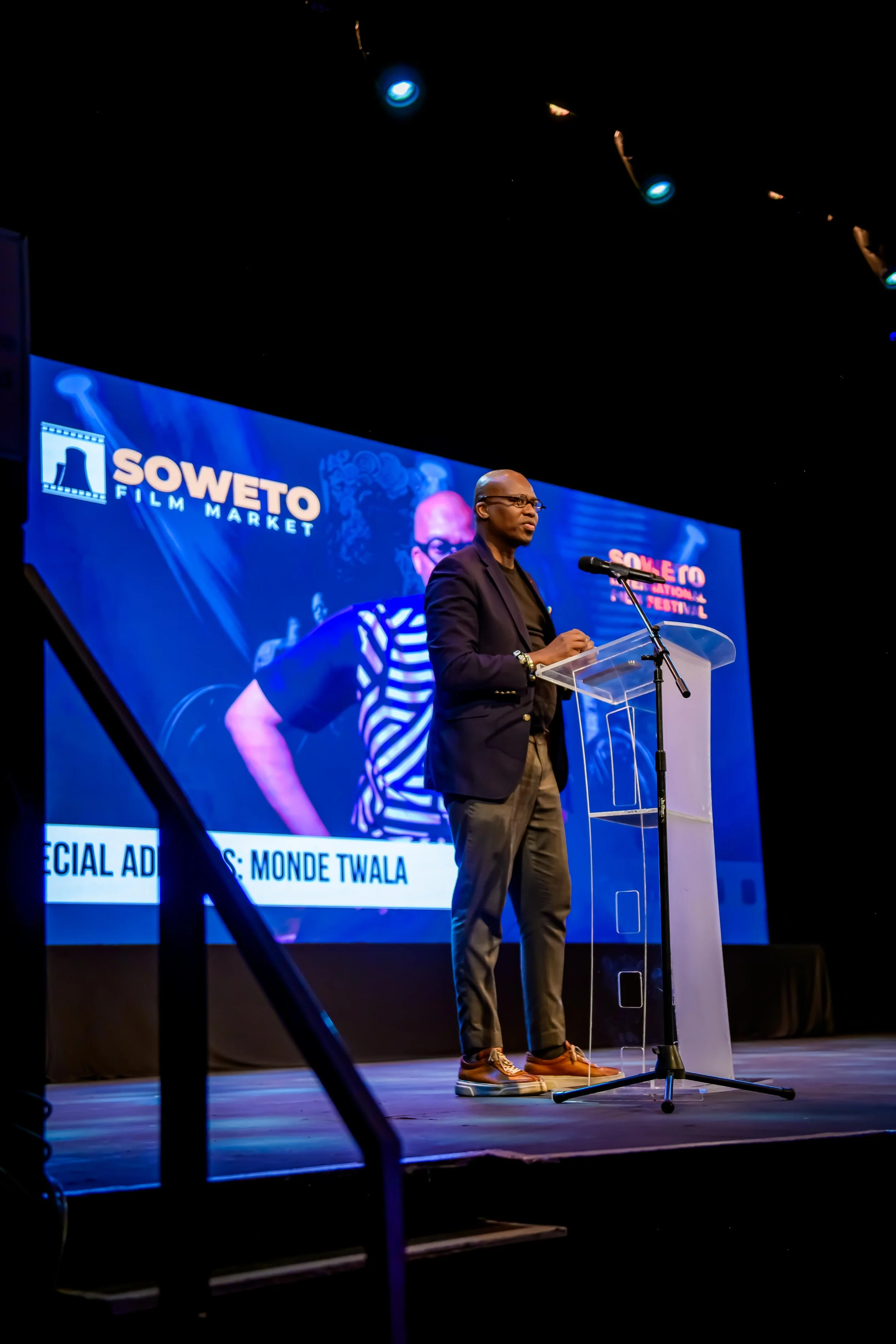 Monde Twala giving a speech on stage at the Soweto Film Market event, standing behind a clear podium with a microphone, with a large screen behind him displaying the event logo and his name, Monde Twala.