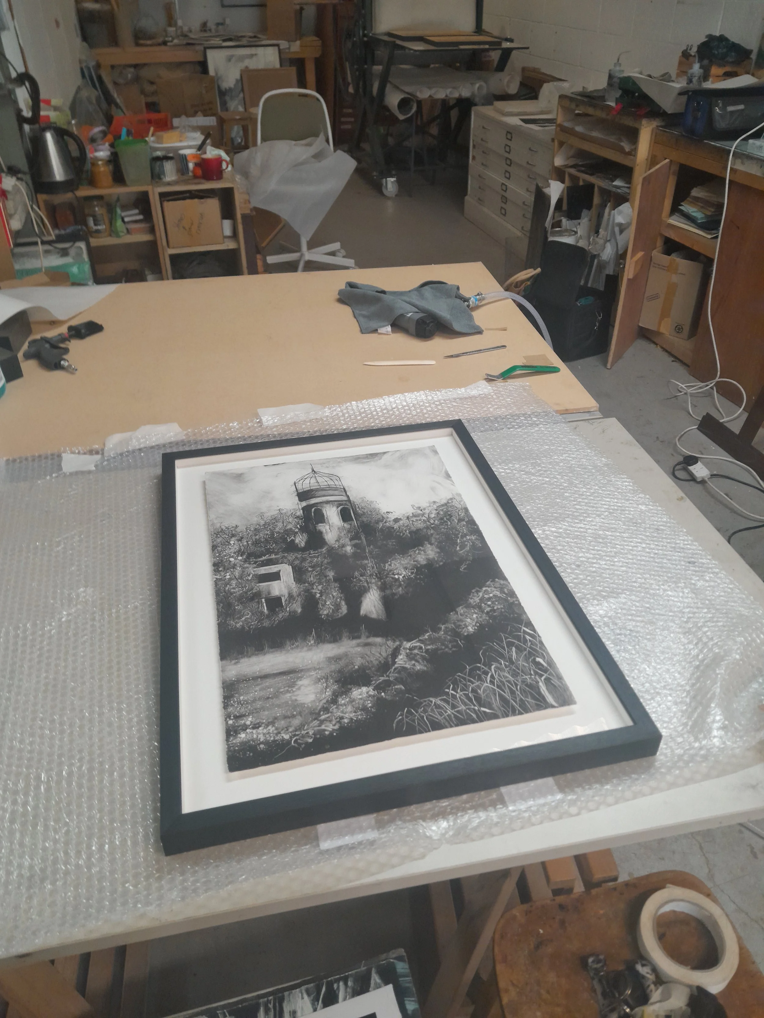 A framed black and white landscape photograph of an old lighthouse on a hill, with trees and a path in the foreground, placed on bubble wrap on a worktable in a cluttered art studio or workshop.