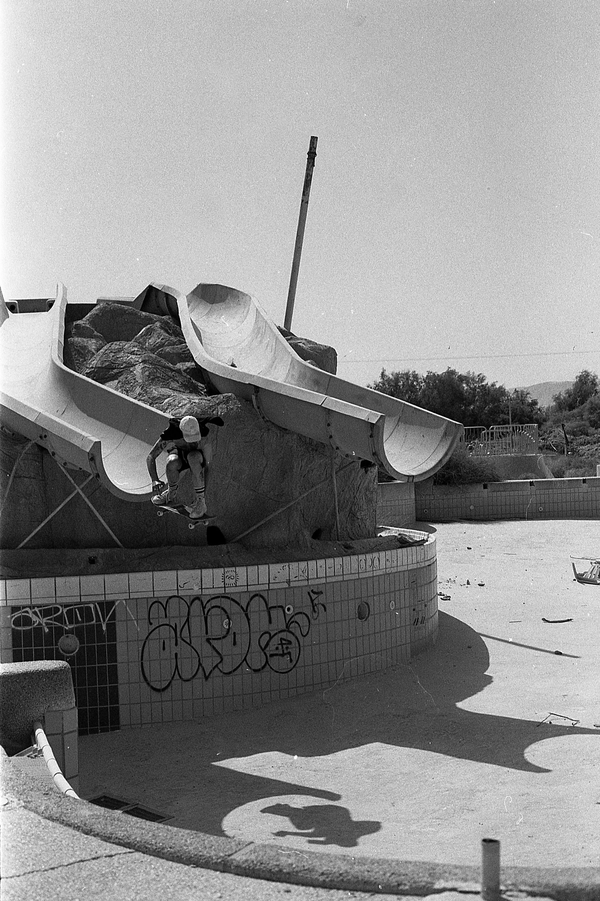 An abandoned outdoor water park with a damaged slide and graffiti on the tiled wall. A person is skateboarding near the slide.
