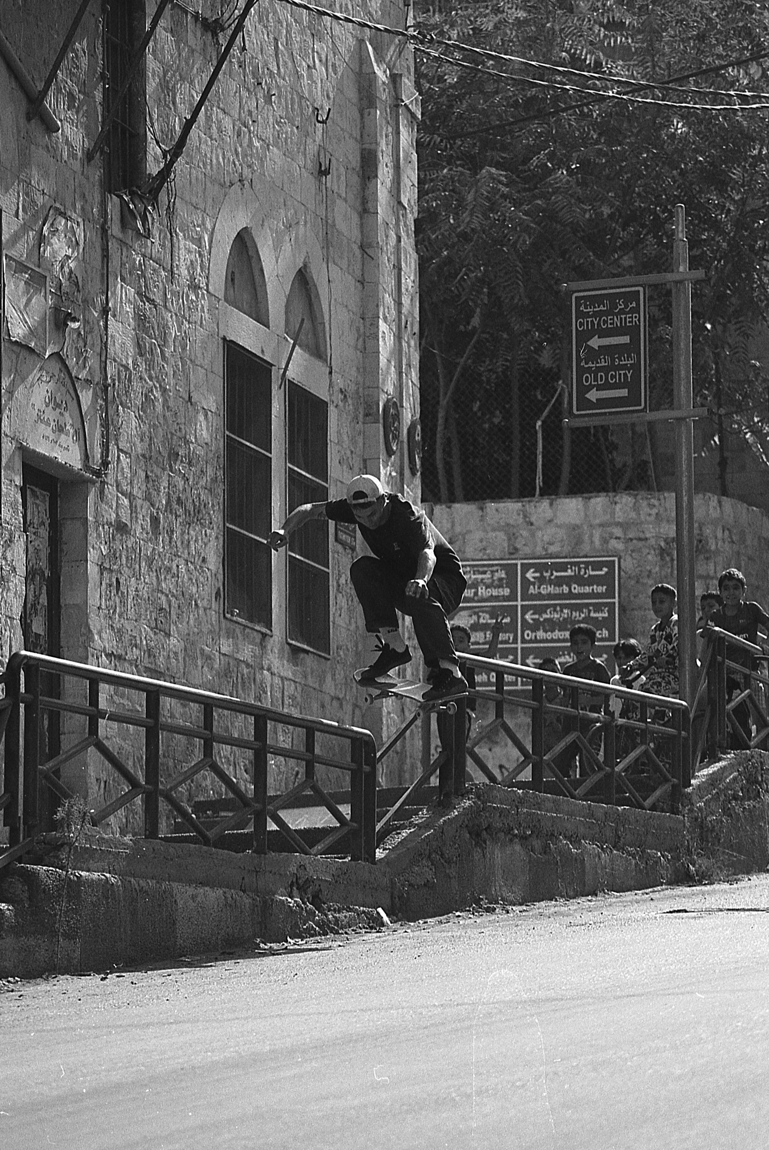 A skateboarder performs a grind on a handrail on a street, with a stone building and a group of children watching in the background.