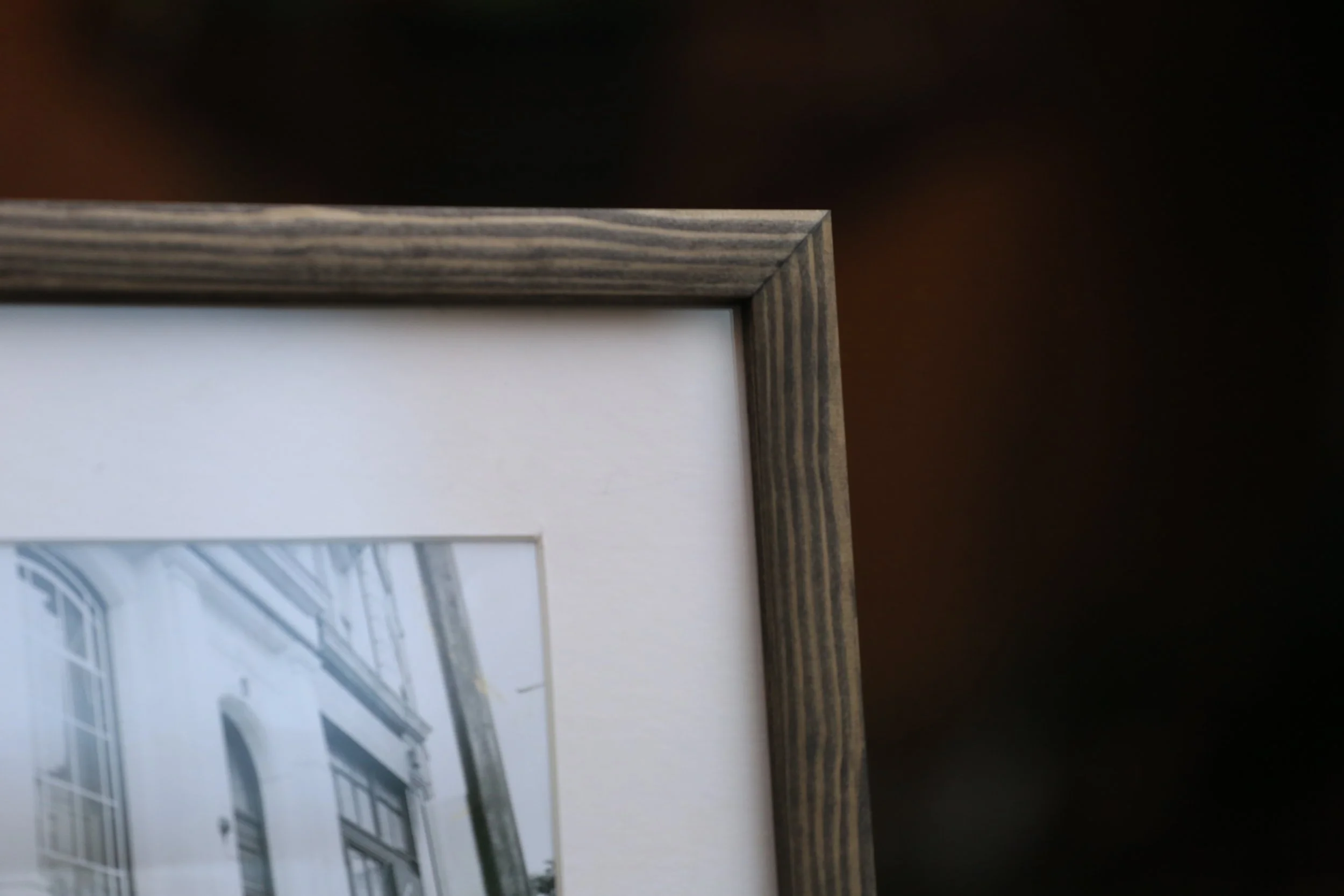 Close-up of the corner of a wooden picture frame surrounding a black-and-white photograph of a building with windows and accordion-style security bars.