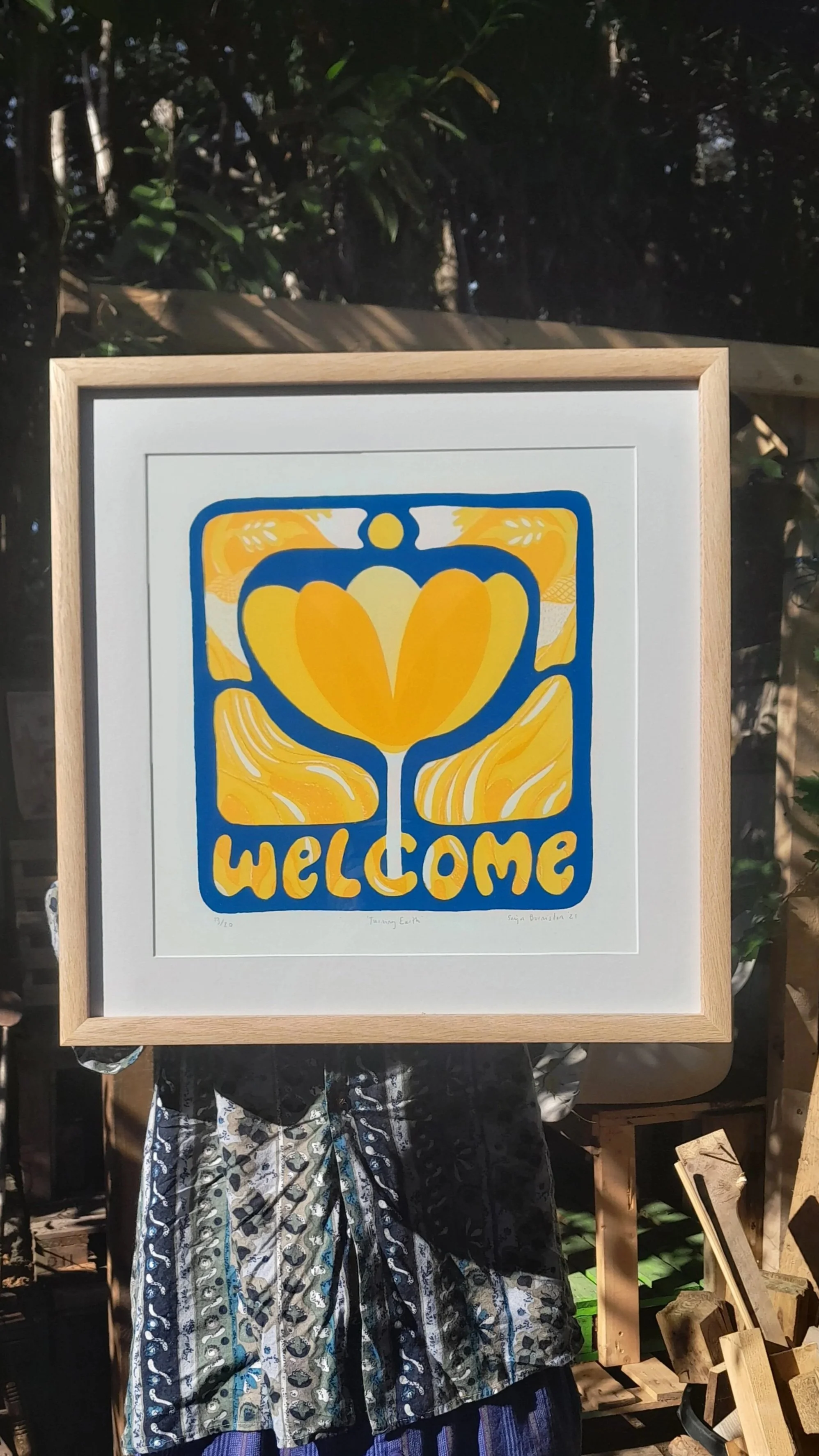 A framed artwork with a yellow, heart-shaped flower and the word 'WELCOME' in bold yellow letters, set against a blue outline, displayed outdoors.