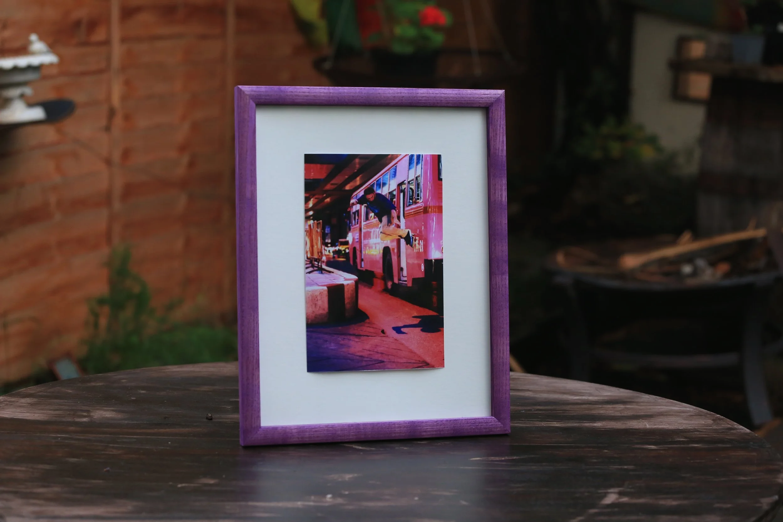 Photograph of a framed picture on a wooden table, showing a person jumping with a skateboard in front of a red and pink tram at a station, outdoors during the daytime.