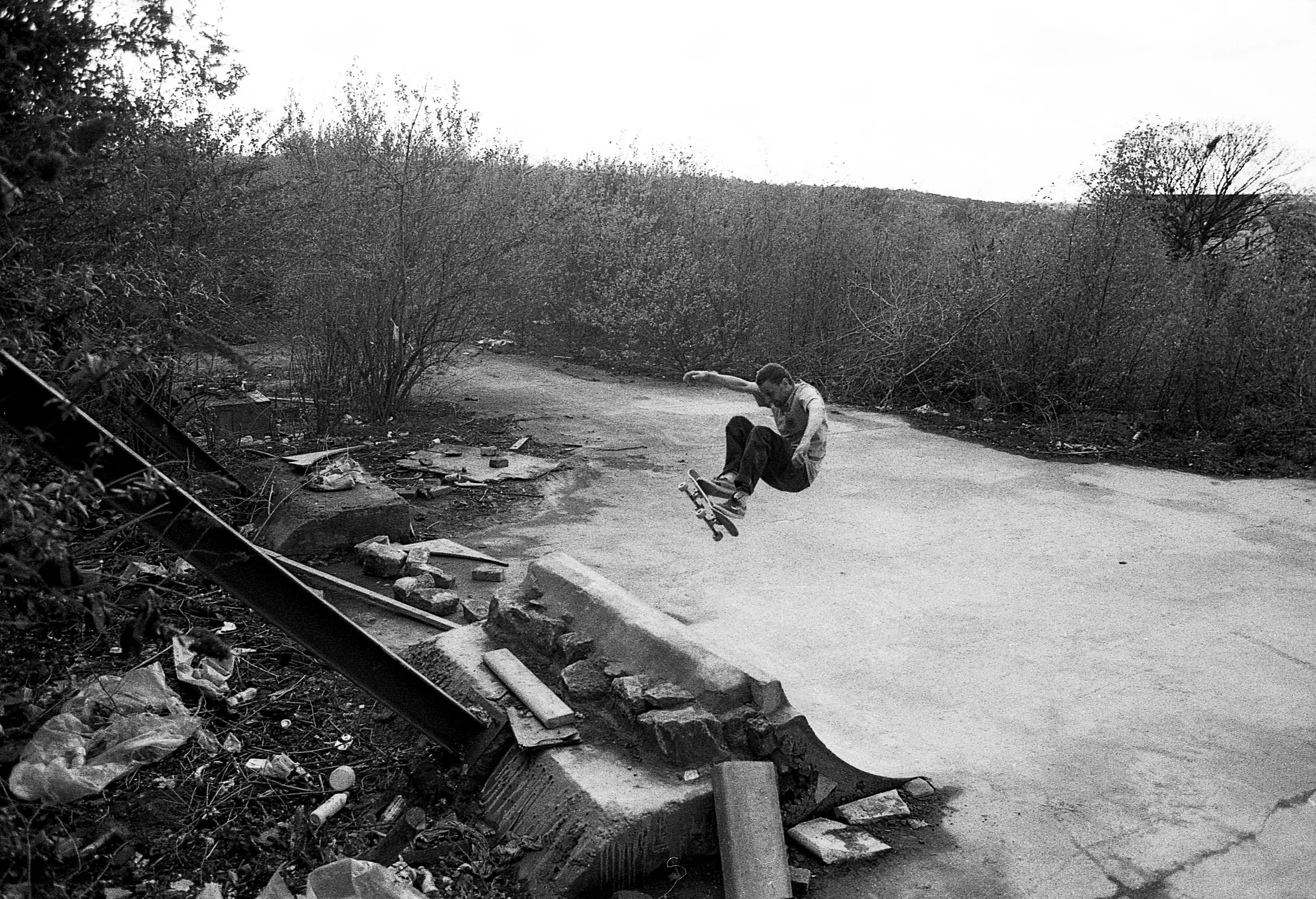A person skateboarding on a road surrounded by trees, with debris and a damaged guardrail on the side.