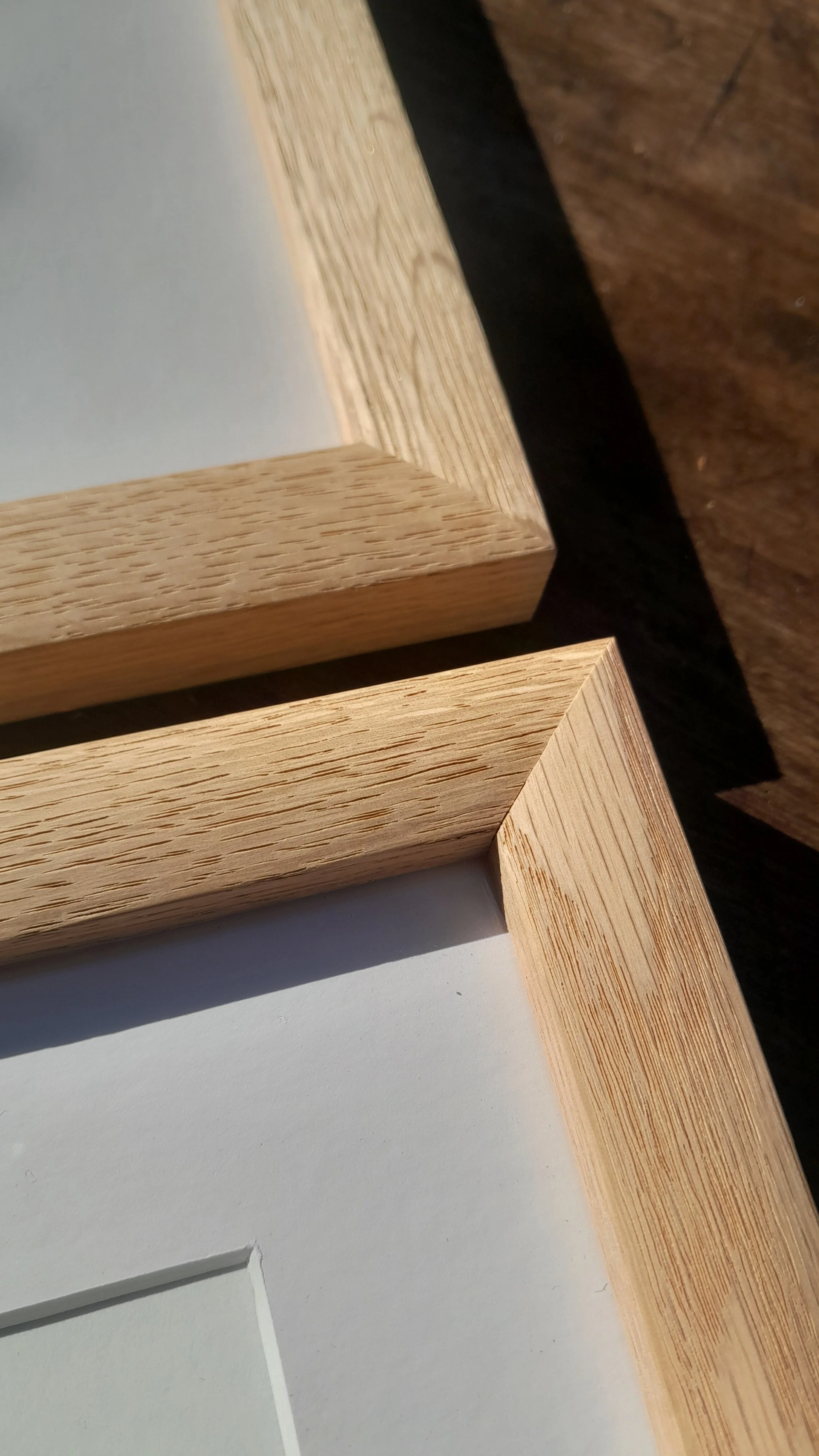 Close-up of the corner of a wooden picture frame with a glass front, showing the wood grain and white mat inside.