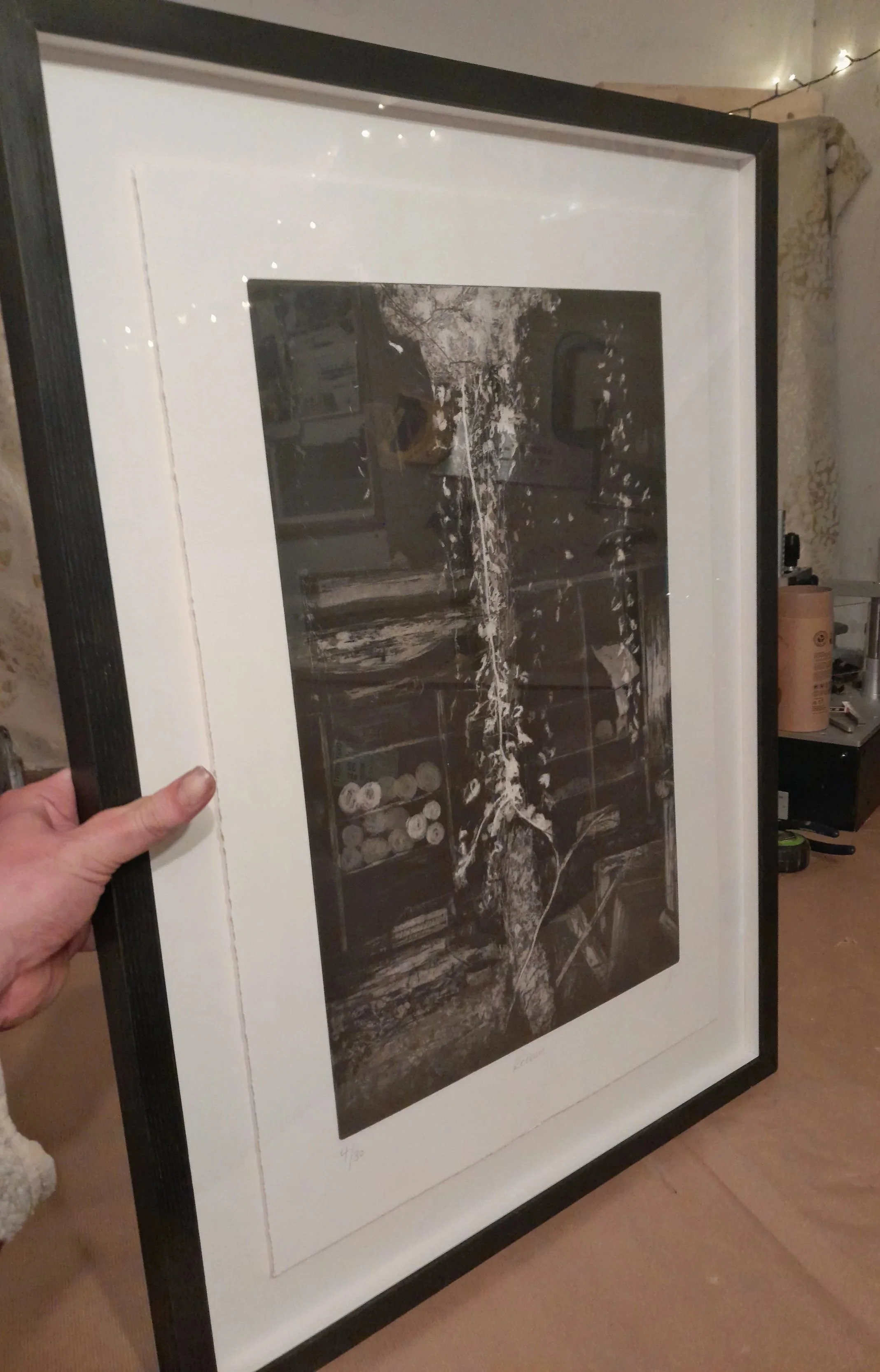 A person holding a framed black and white photograph with a white border, showing some signature and edition number at the bottom, in an indoor setting with string lights and assorted items in the background.