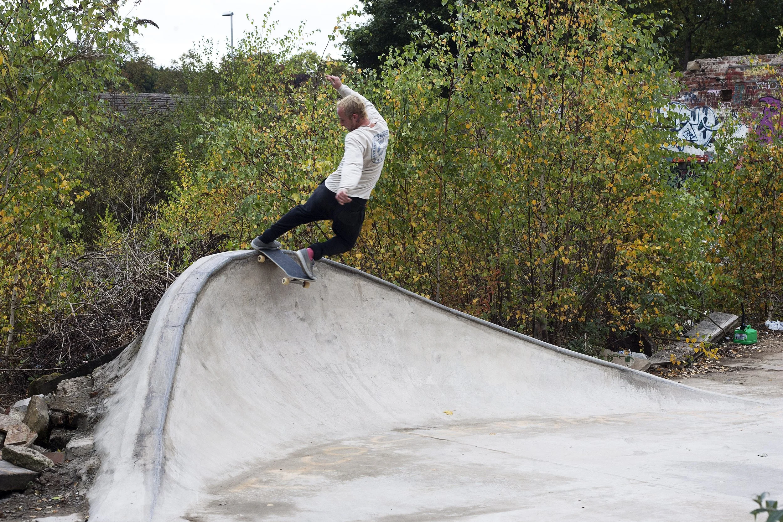 A skateboarder is performing a trick on the edge of a concrete skate park ramp with trees and graffiti in the background.