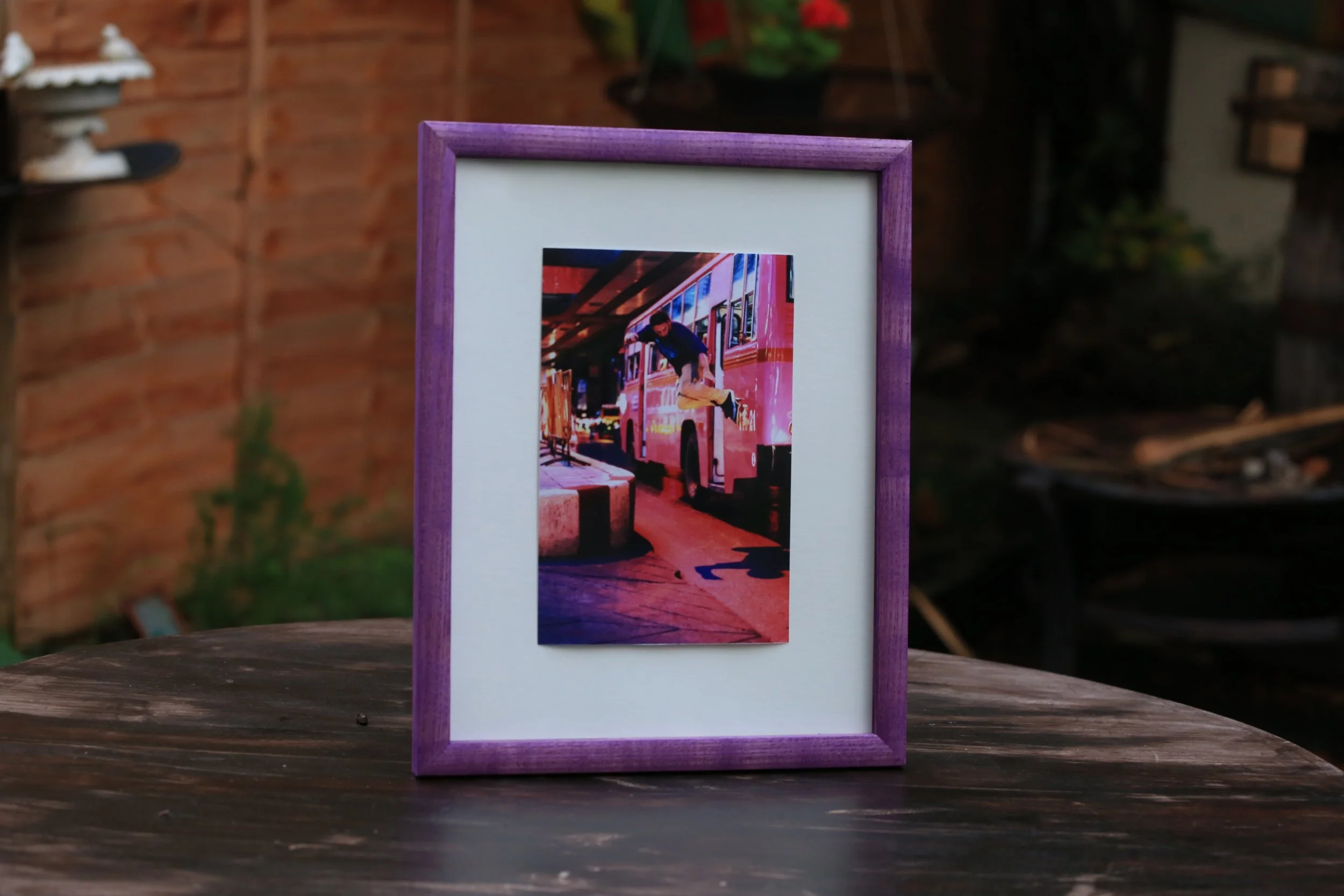A framed photo of a man jumping onto a pink bus inside a building, with a street and other buses visible in the background.