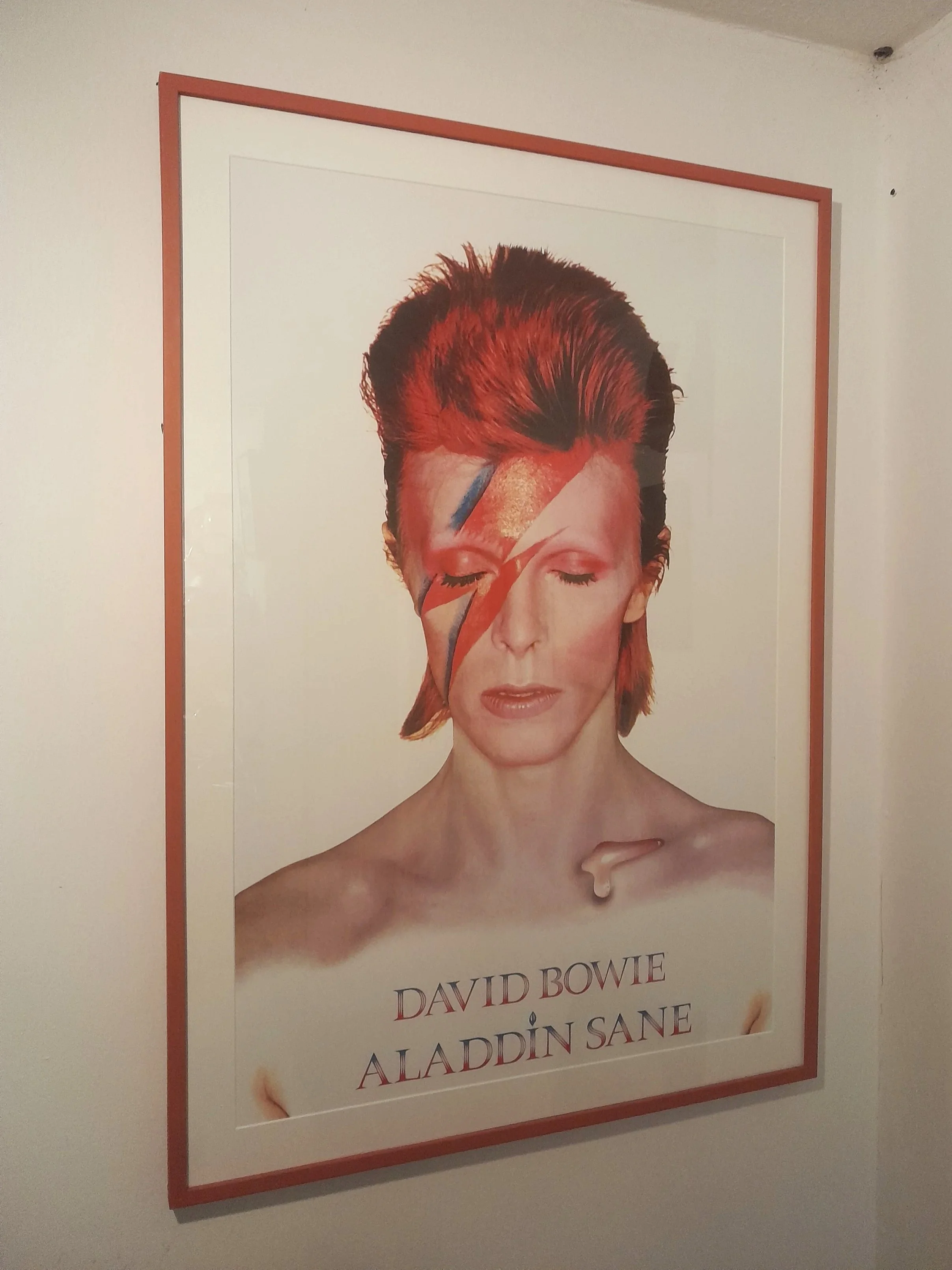 Poster of David Bowie with his eyes closed, red hair, and a lightning bolt painted on his face, referencing his Ziggy Stardust era.
