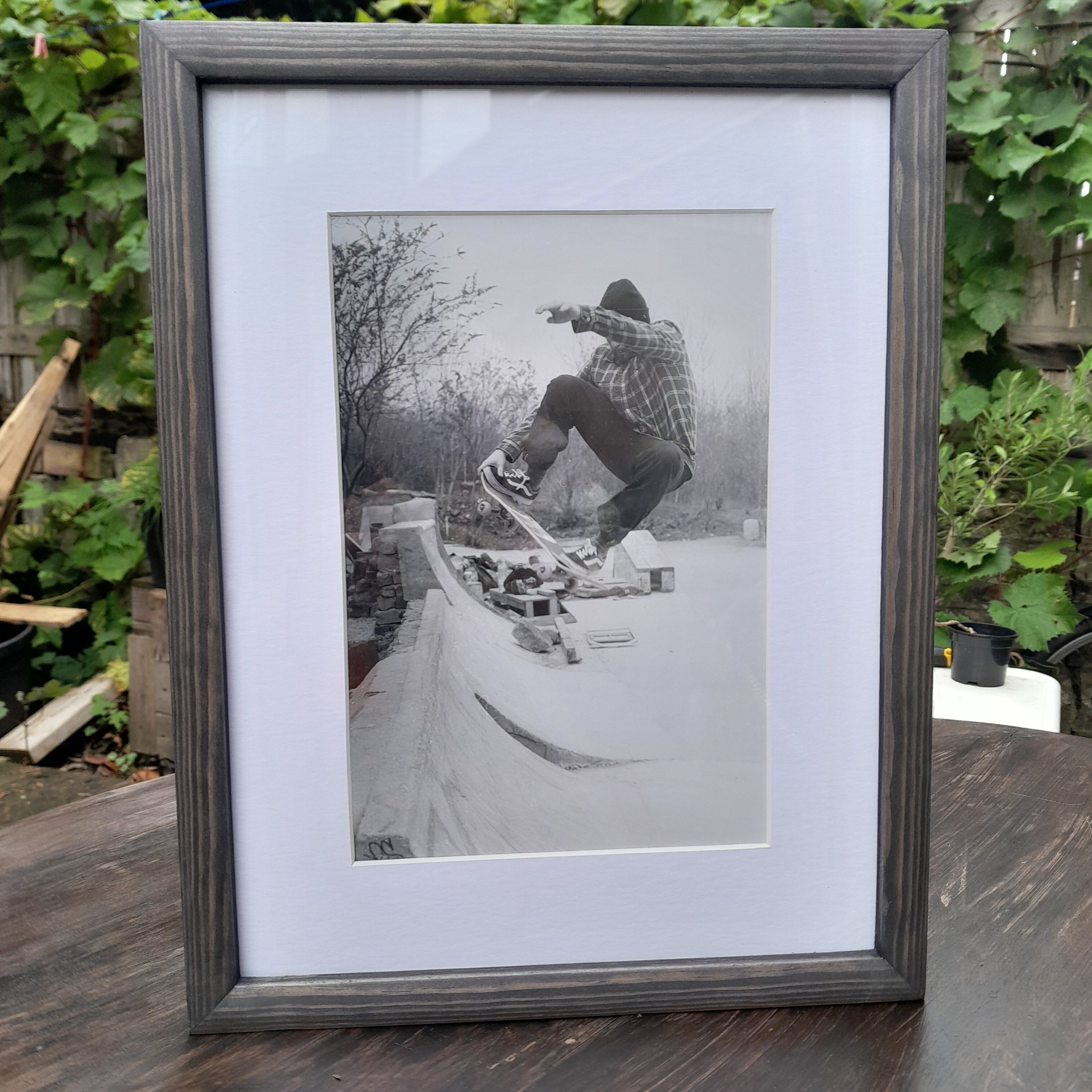 Black and white photo of a skateboarder performing a trick on a ledge outdoors, with trees and a clear sky in the background.