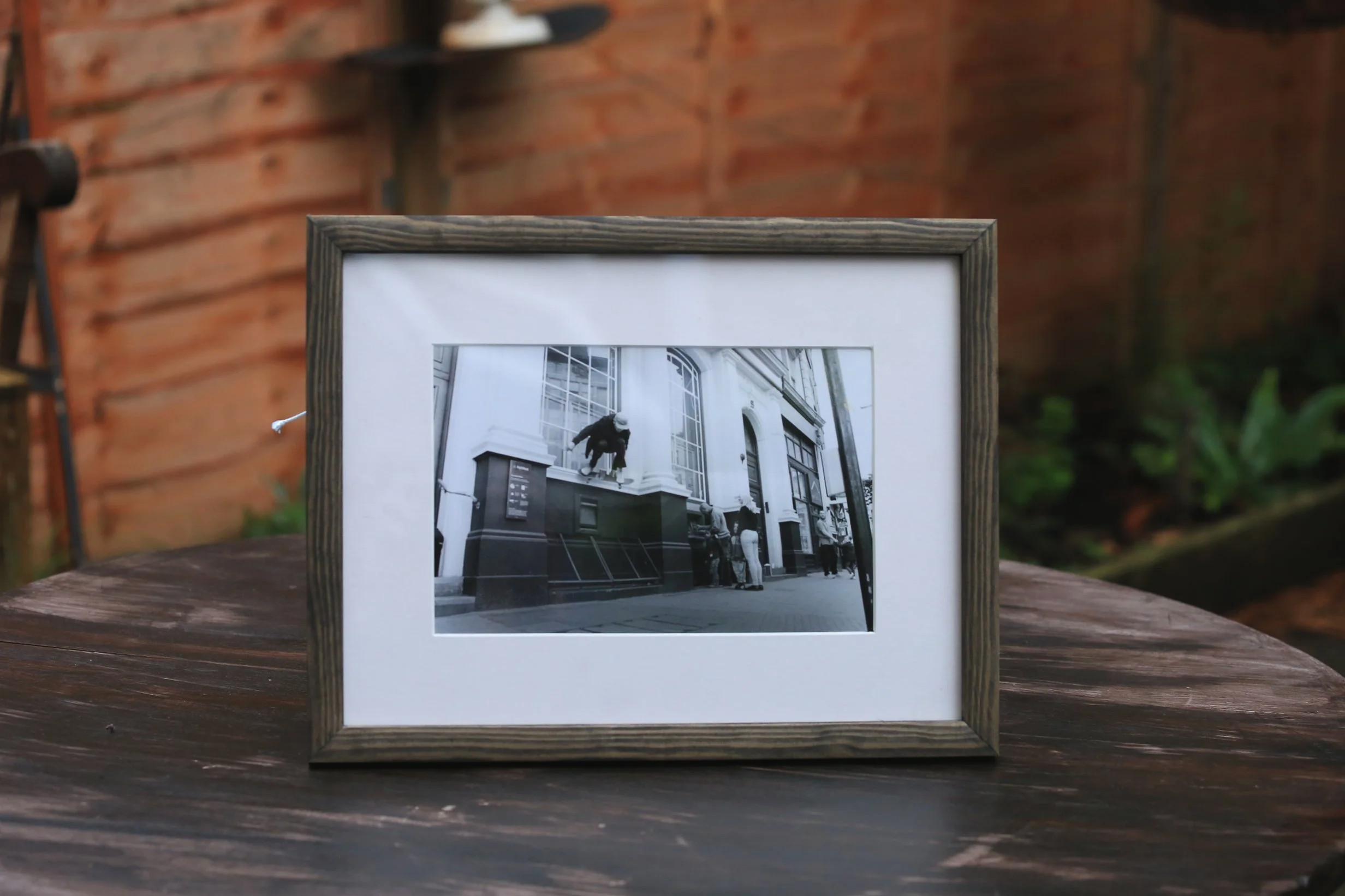 Black and white photograph in a wooden frame showing a city street scene with a person jumping off a building facade and a group of people standing on the sidewalk in front of a tall building.