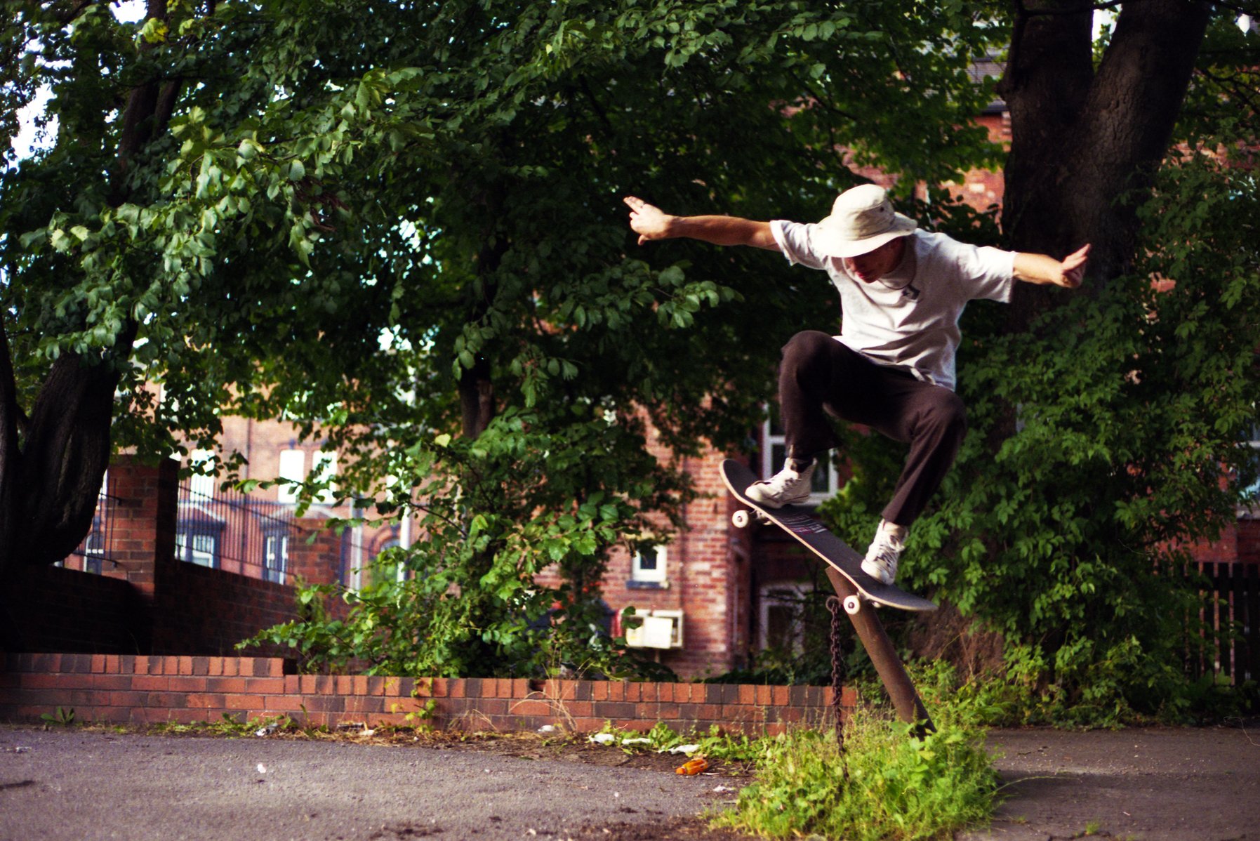 Skateboarder performing a trick on a broken and chained skateboard in a park with green trees and brick buildings in the background.