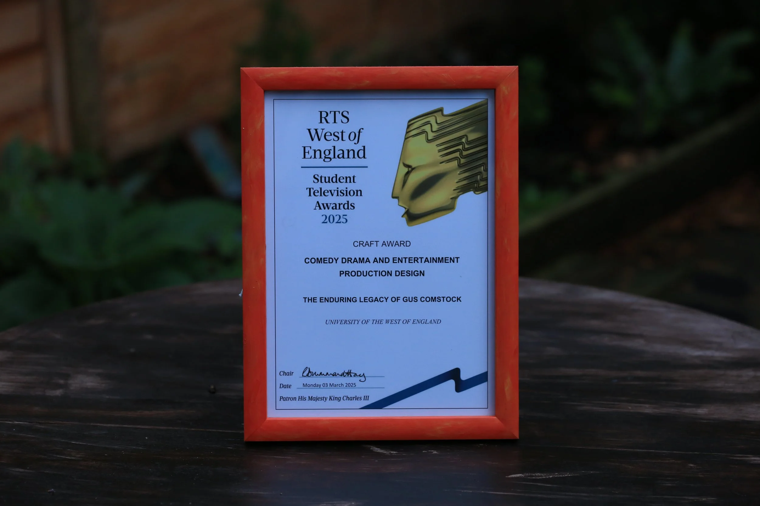 A framed award certificate from the RTS West of England 2025 Student Television Awards for a craft award in production design, for a project titled 'The Enduring Legacy of Gus Comstock' from the University of the West of England. The certificate is placed on a dark wooden surface with a blurred outdoor background.